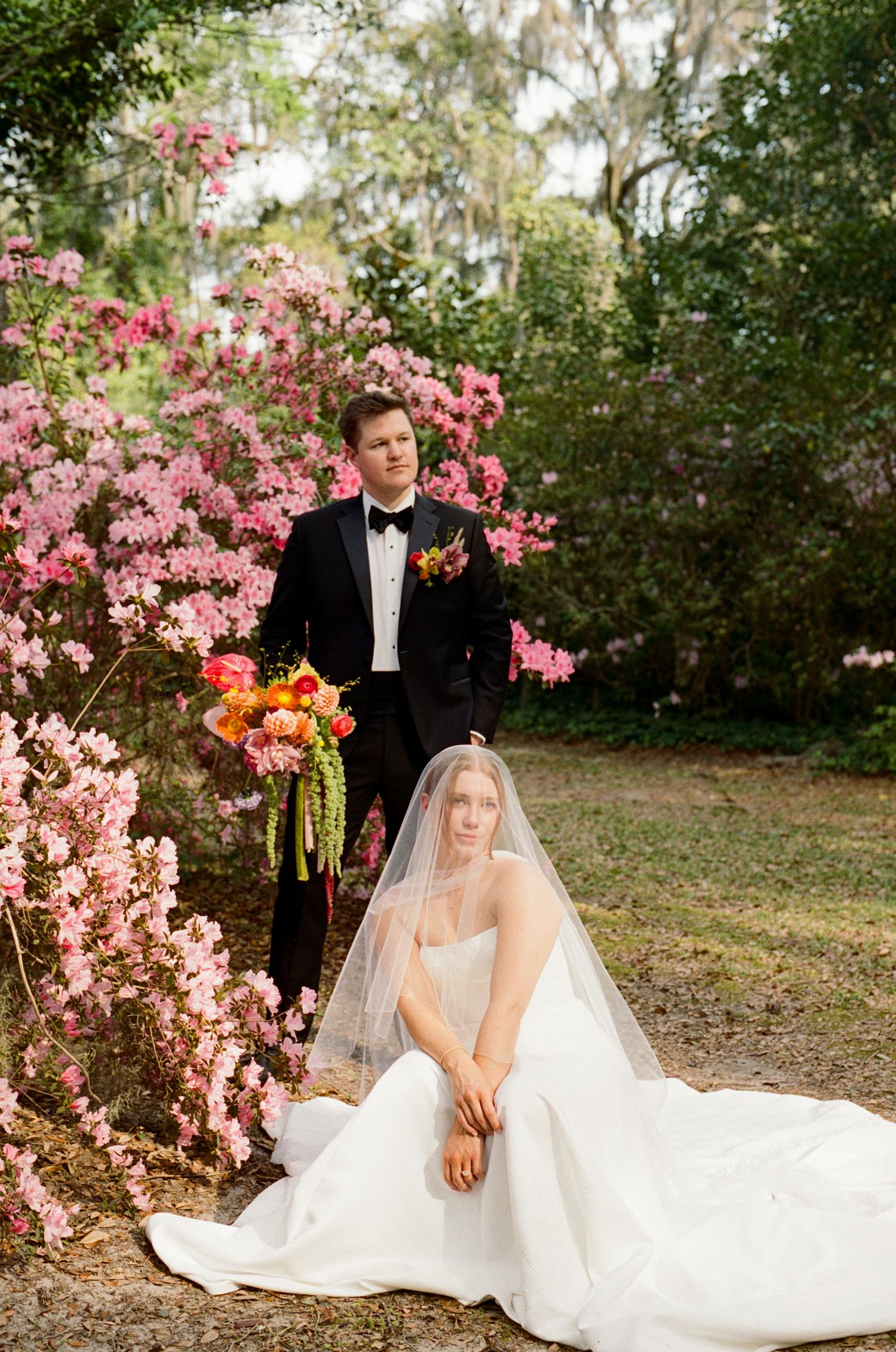 Bride in white gown with veil sits before groom in black tuxedo amid blooming pink azaleas outdoors.