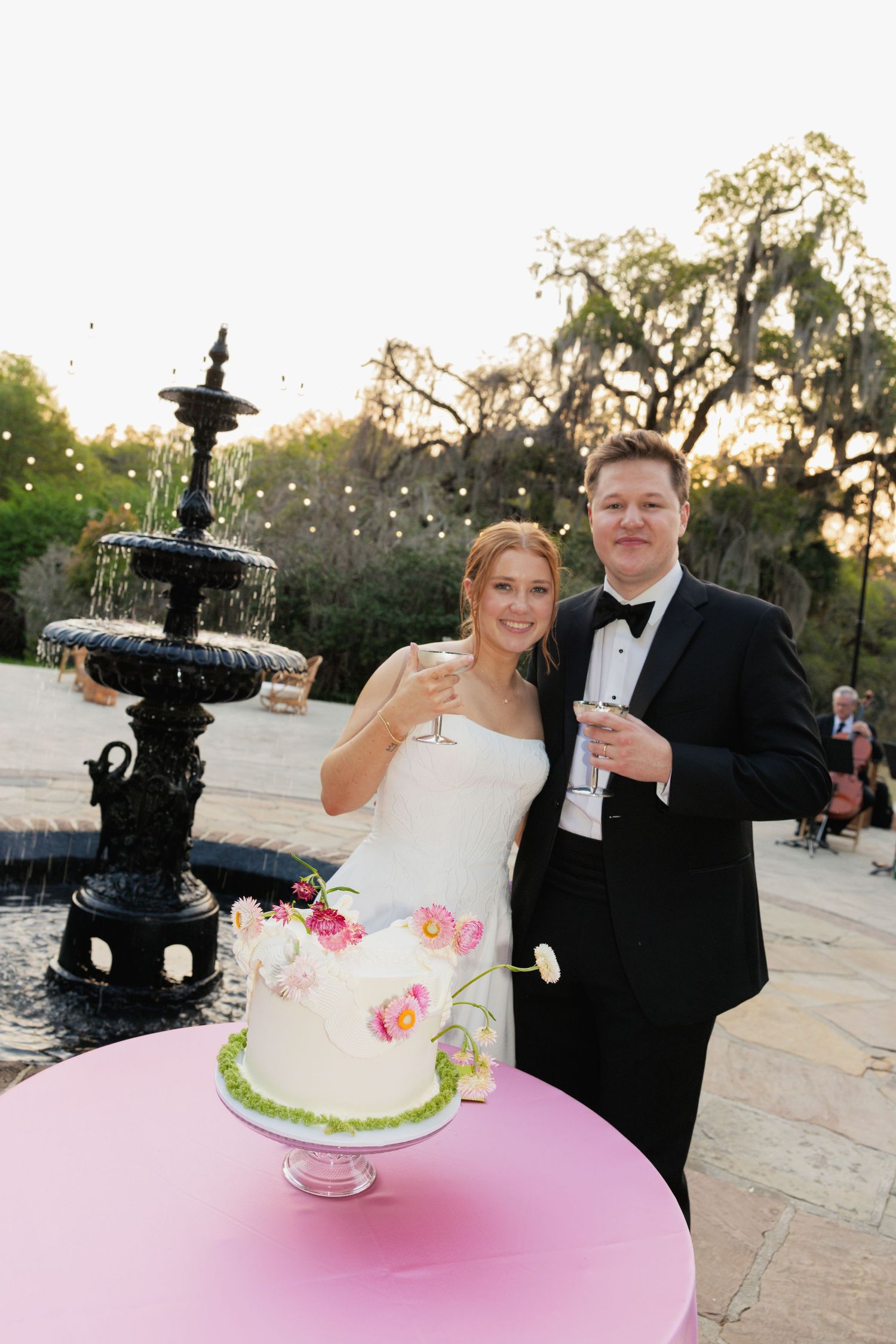 Bride and groom toasting with champagne beside a white floral wedding cake and fountain at outdoor reception.