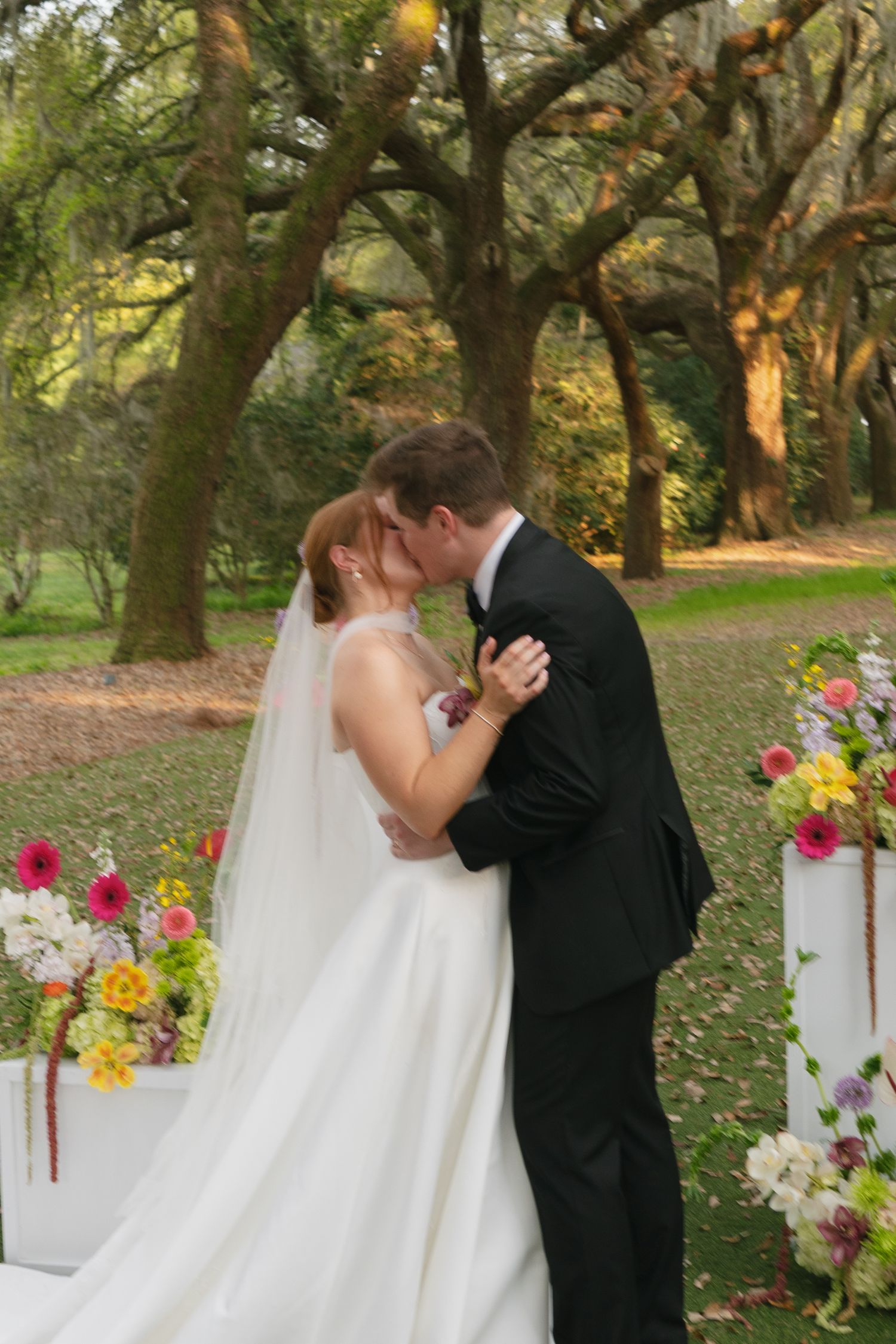 Bride and groom share first kiss at outdoor wedding ceremony surrounded by colorful floral arrangements and moss-draped oak trees.