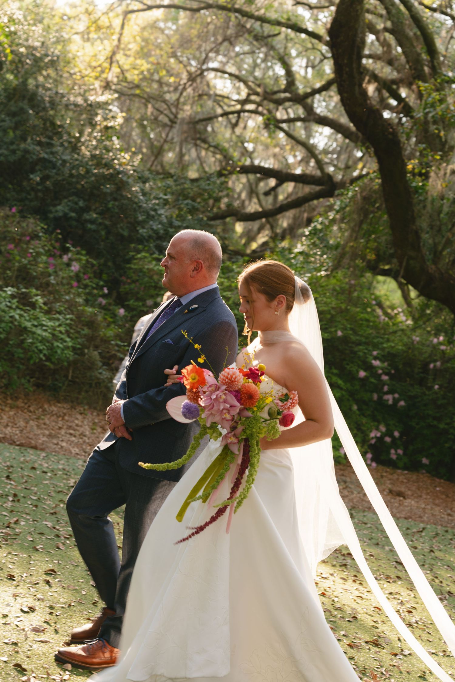 Bride in white gown holding colorful bouquet stands with groom in navy suit in a sunlit garden setting.