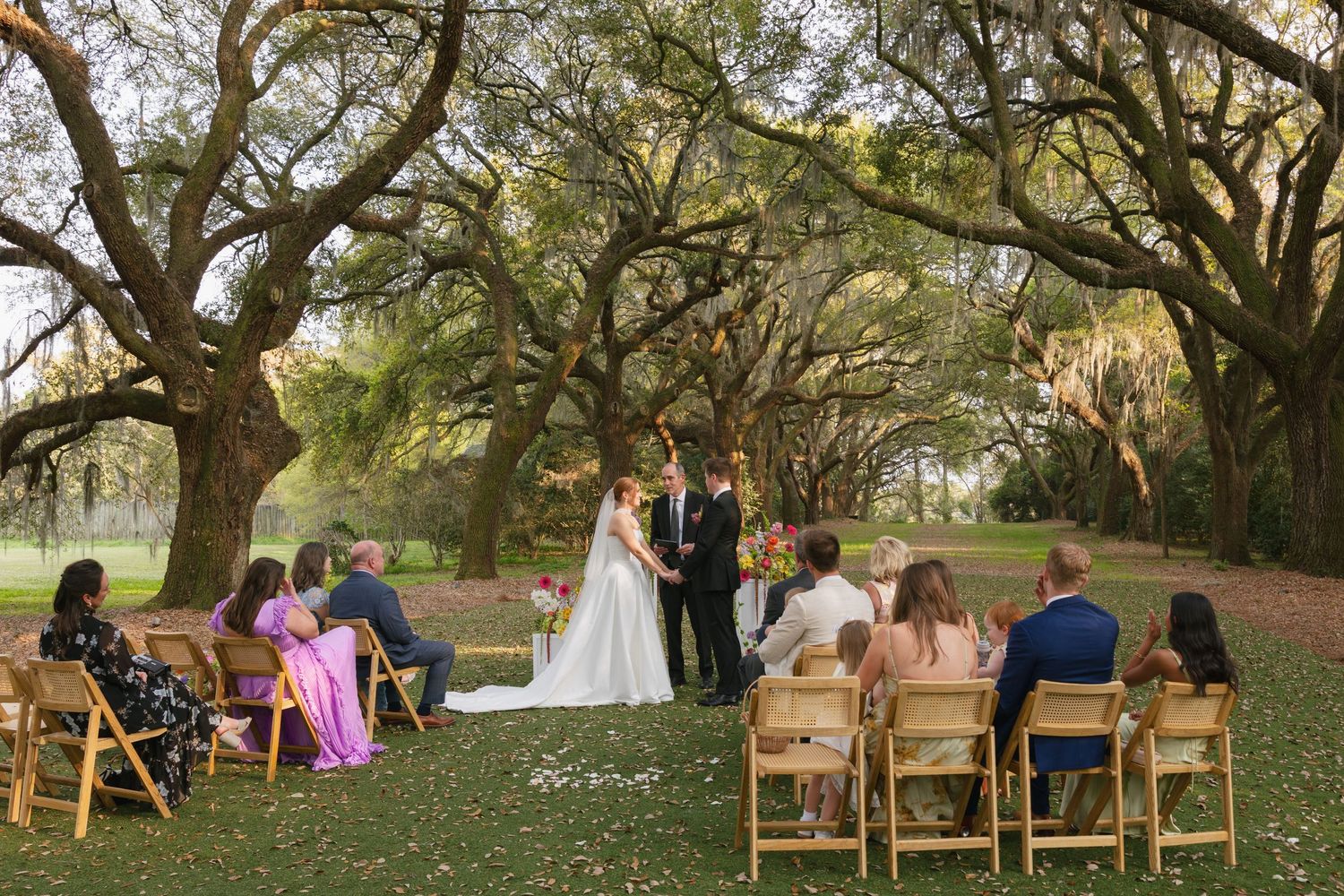 Outdoor wedding ceremony under oak trees with bride and groom exchanging vows before small group of seated guests on lush green lawn.
