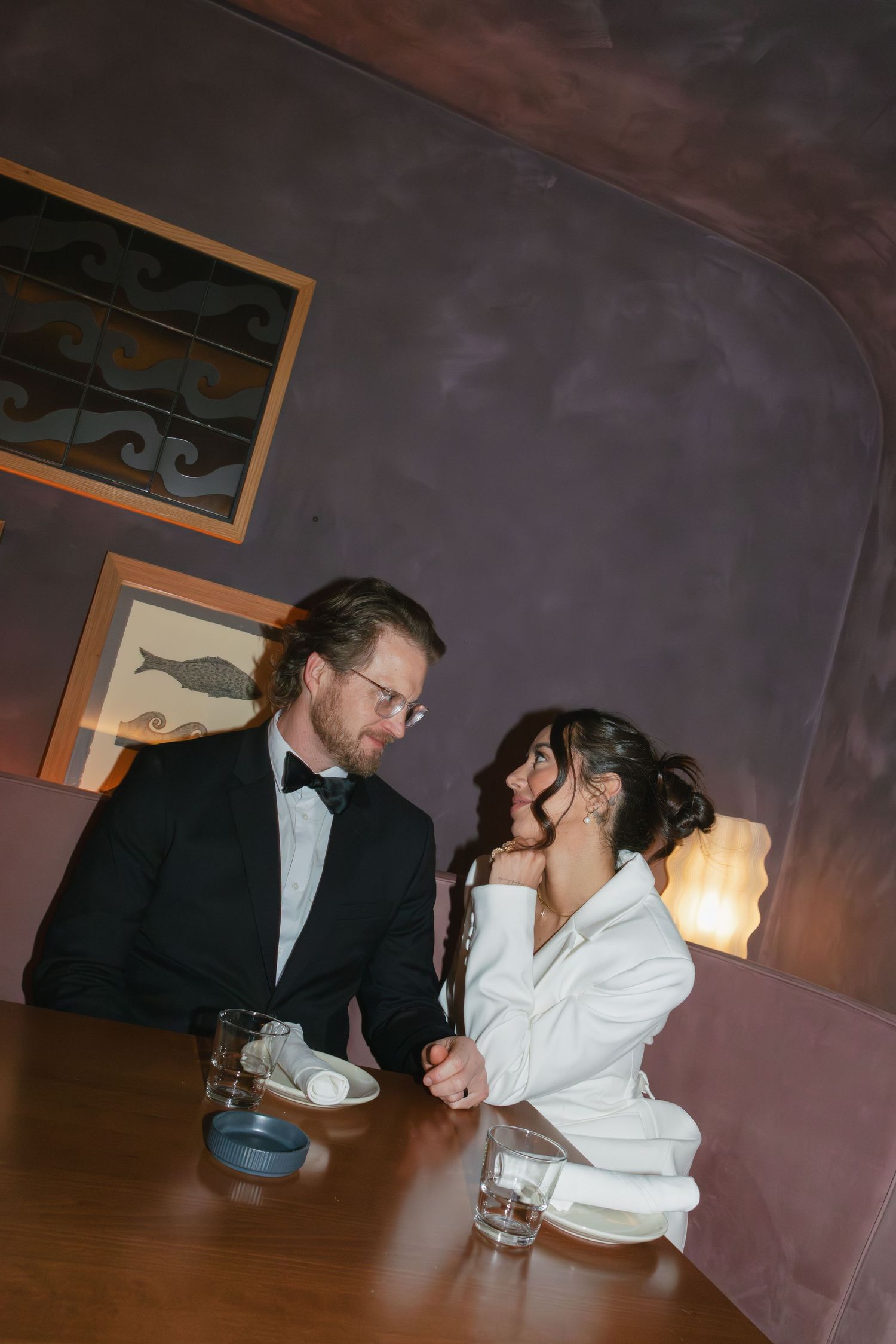 Elegant couple in formal attire sharing an intimate moment at a restaurant table with warm ambient lighting.