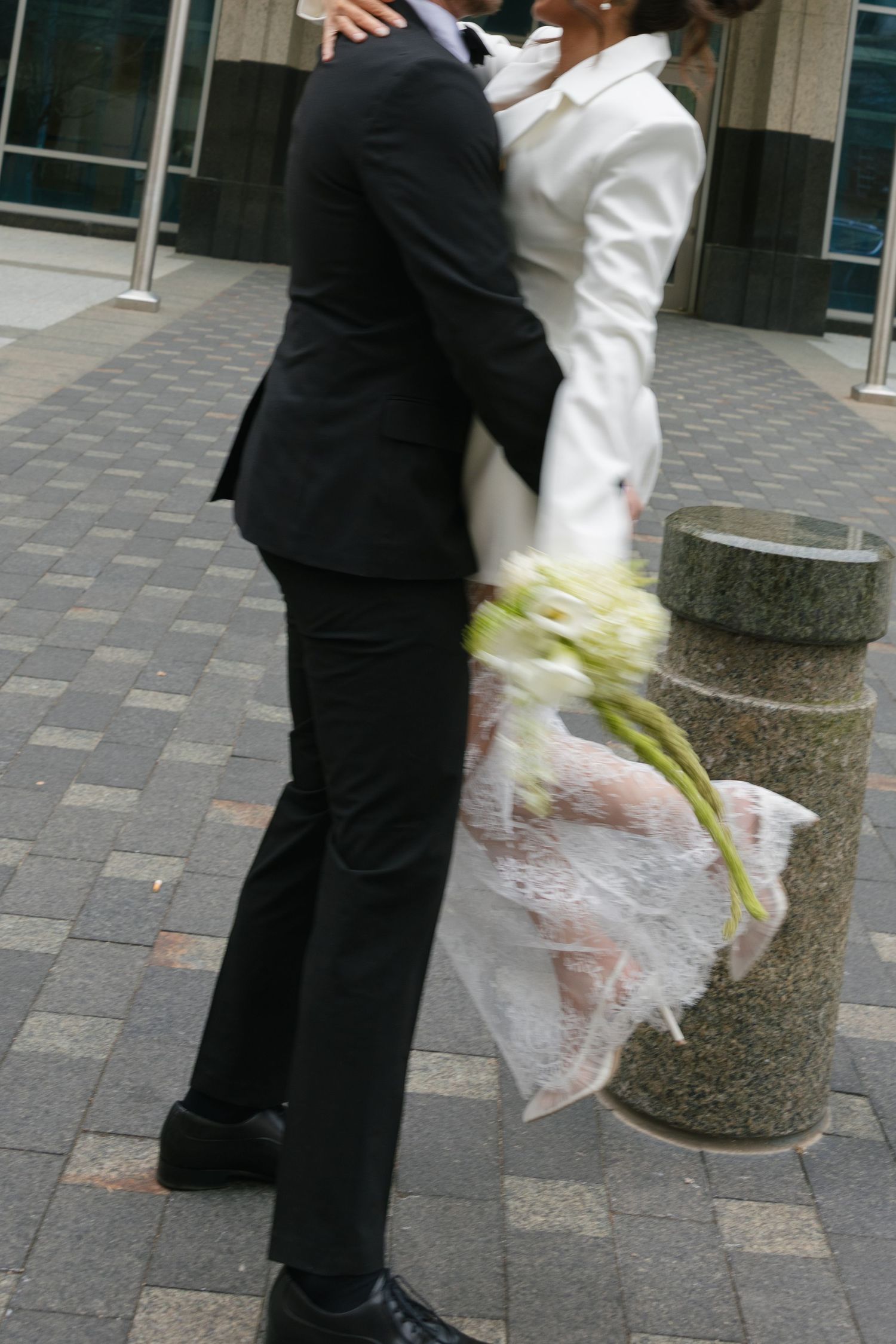 A couple embraces outdoors in wedding attire, the bride holding white flowers in a white suit, groom in a black tuxedo.