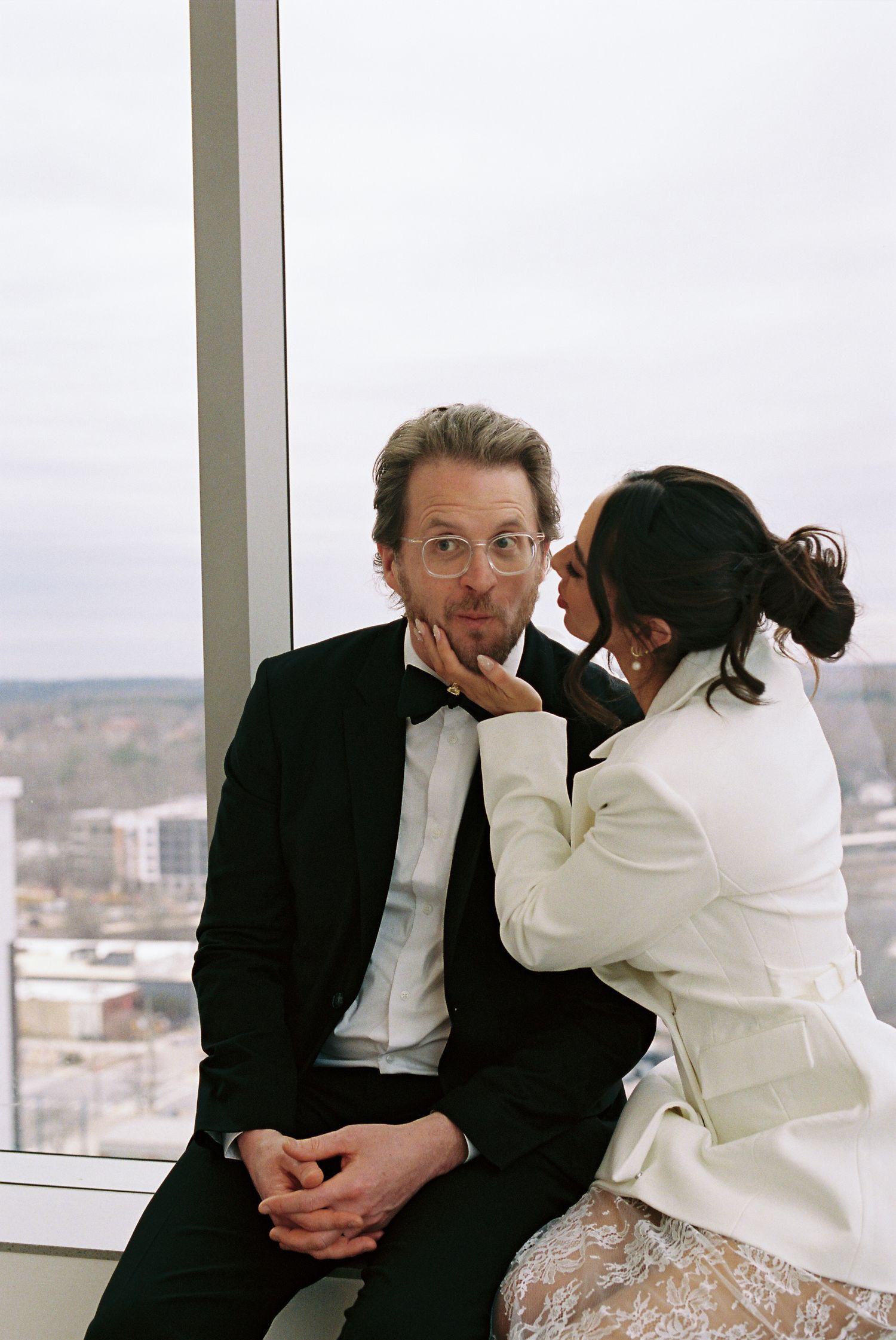 Couple in formal wedding attire sitting by a large window, groom in black tuxedo, bride in white jacket whispering.