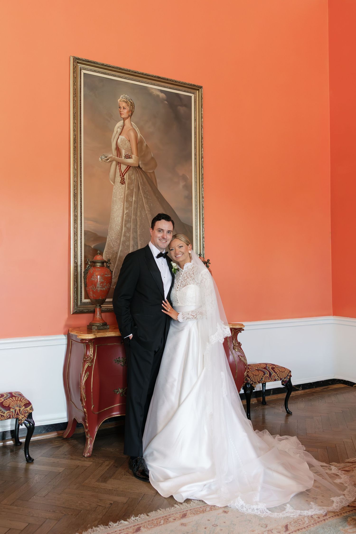 Bride and groom posing elegantly in a coral-walled room with a vintage portrait painting and ornate red console table.