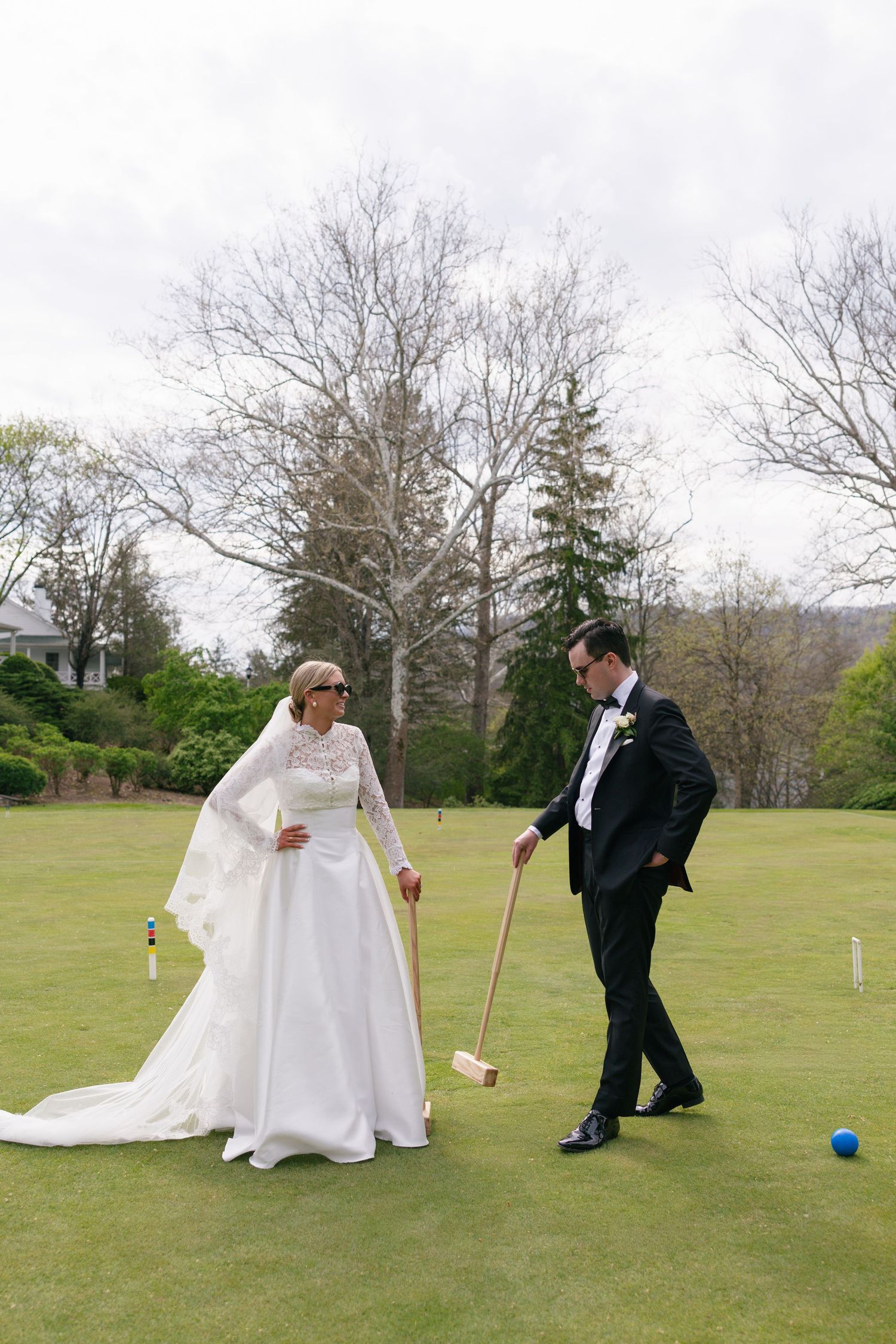 Bride in white gown plays croquet with groom in tuxedo on lush green lawn during outdoor wedding celebration.