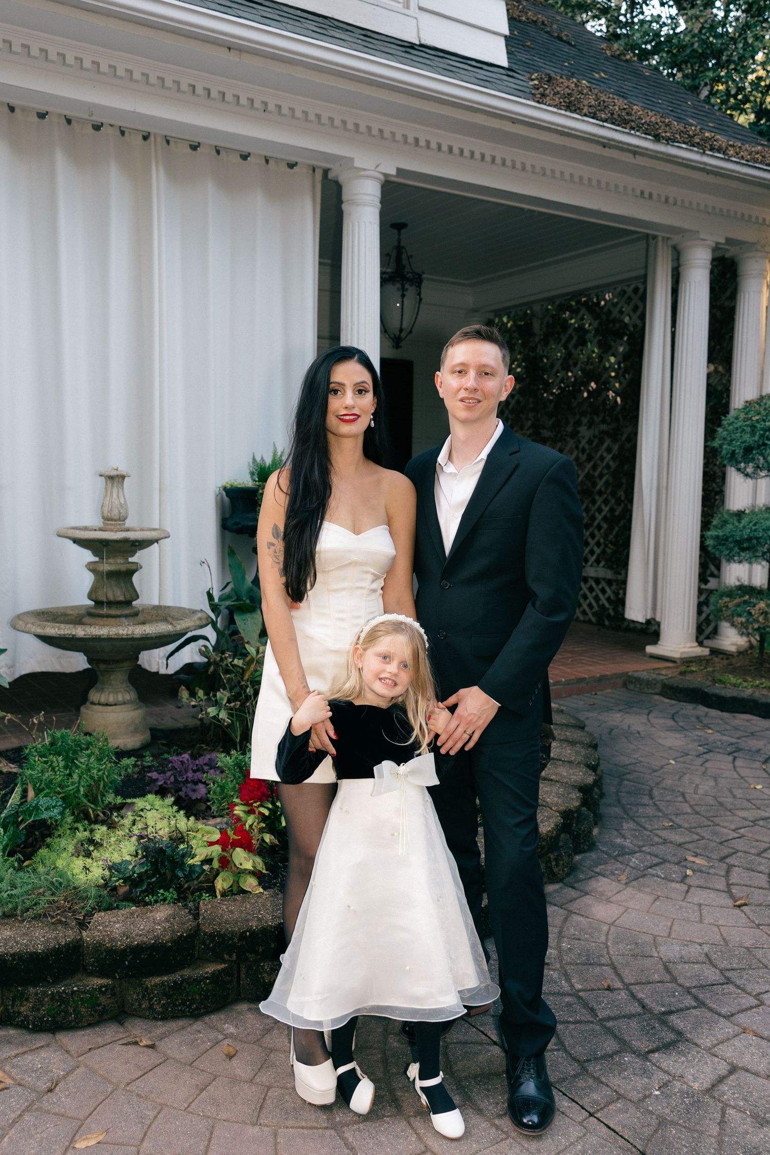 Smiling family of three in formal attire poses on a brick patio beside a stone fountain and garden at a white columned porch.