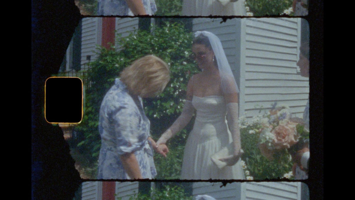 Bride in a white gown and veil holds hands with a woman in a blue patterned top outdoors near green shrubs and pink flowers.