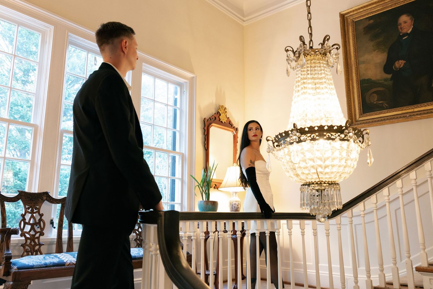 Man in a dark suit faces a woman in a black dress and gloves on an elegant staircase landing beneath a crystal chandelier.