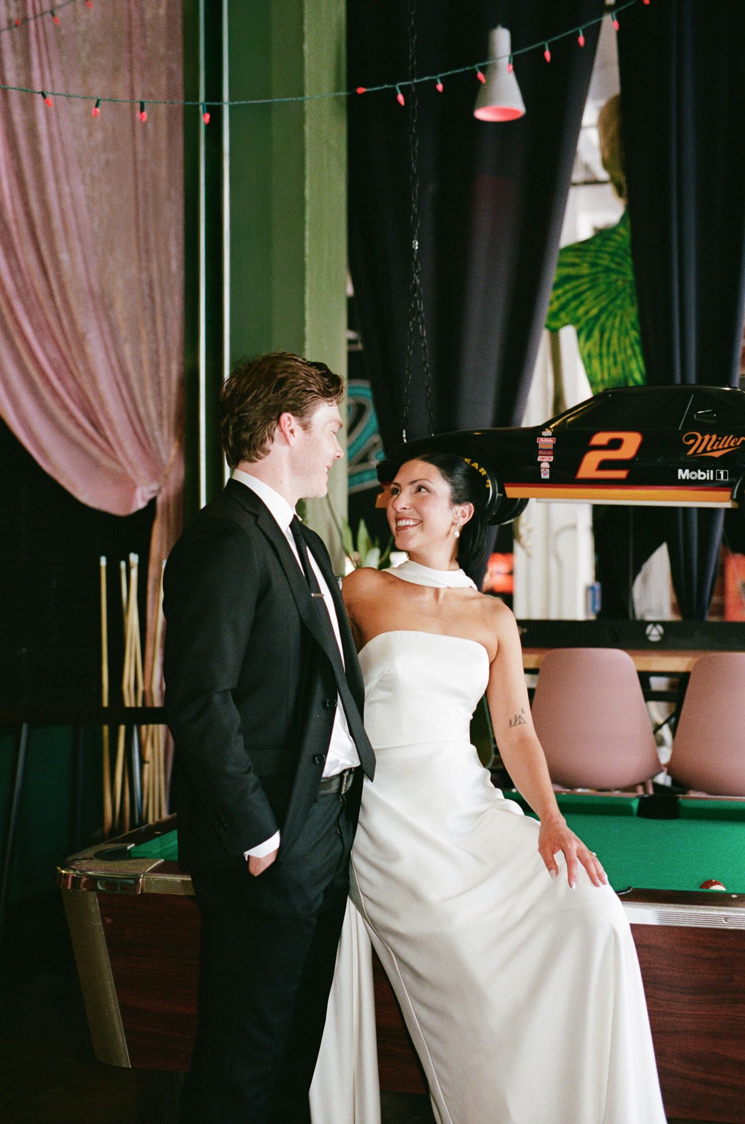 Bride in a strapless white gown and groom in a black suit pose beside a green pool table with a racing car display behind them.
