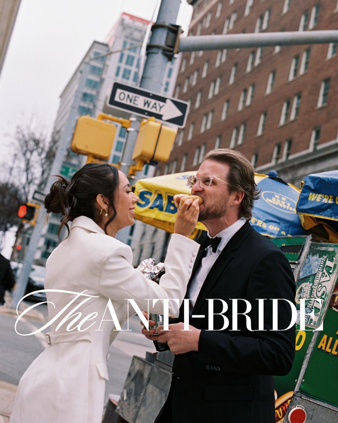 Bride in white coat feeds groom in tuxedo a hot dog on a NYC street corner, with One Way sign above.