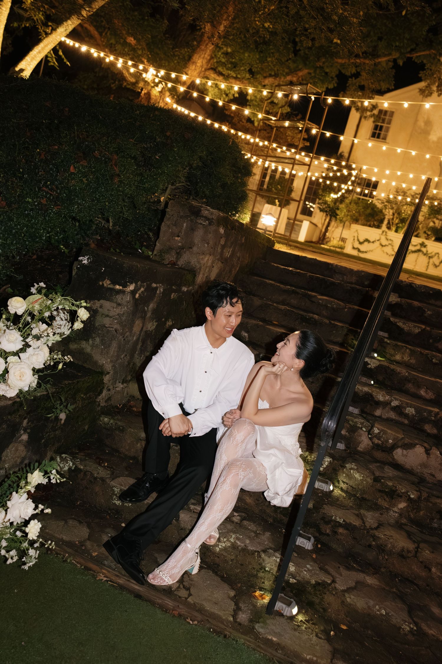A romantic evening scene under string lights with a couple sharing a tender moment on stone steps.