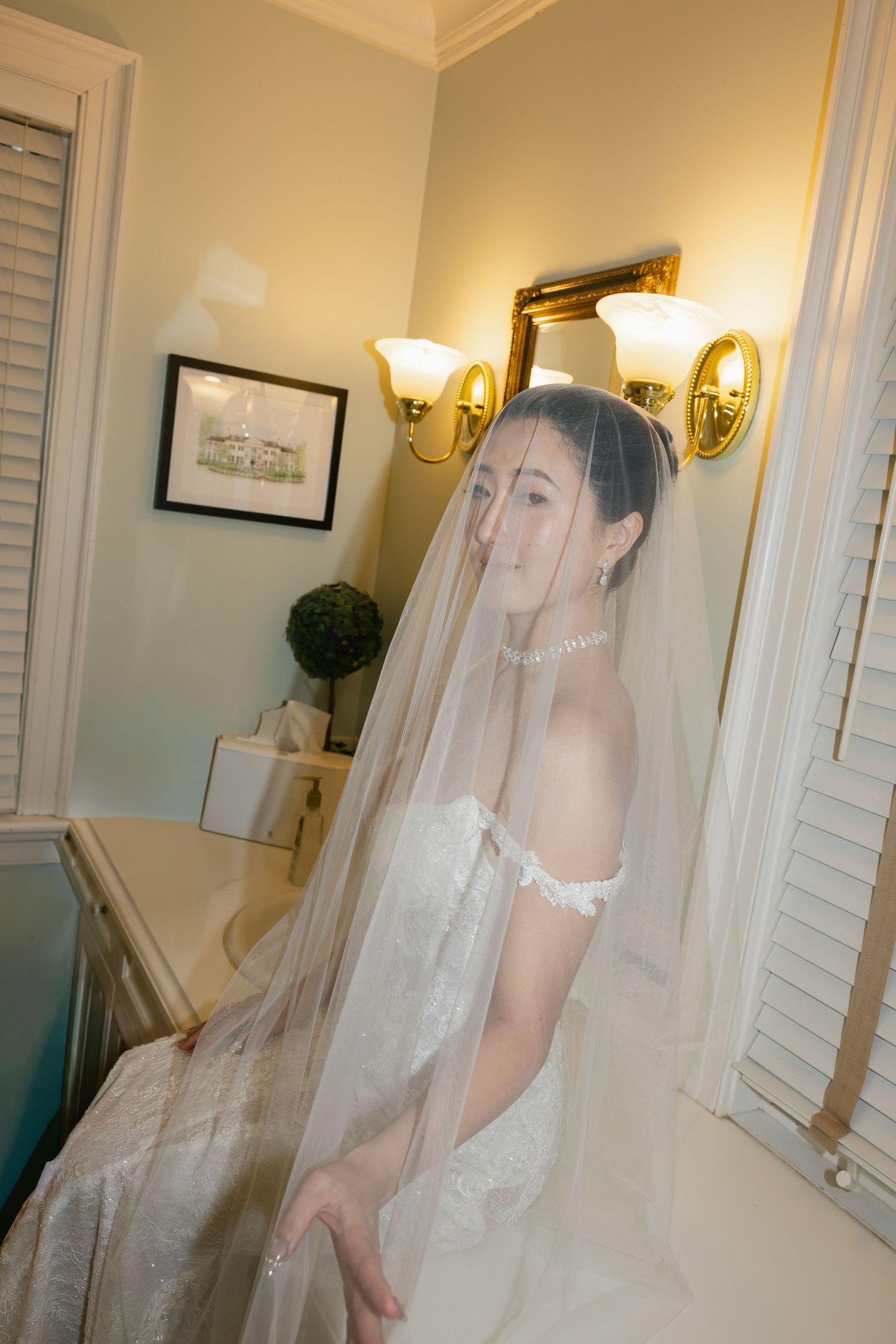 A bride in a white wedding dress and veil stands in an elegant room with wall sconces and classic decor.
