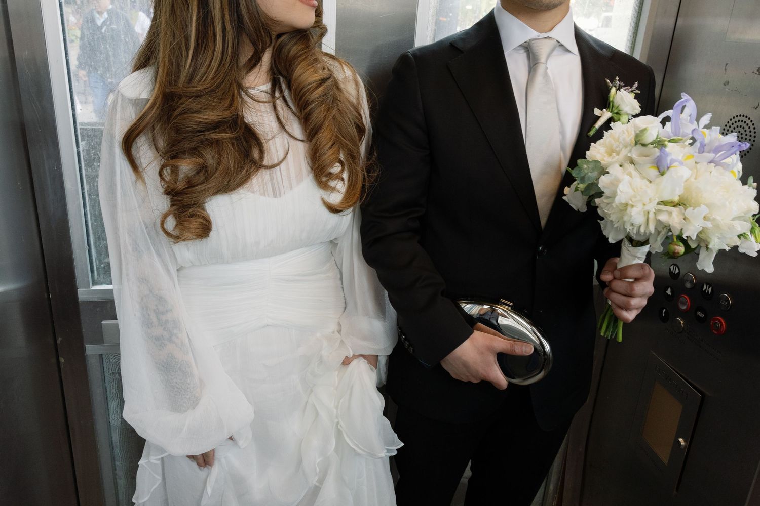 A couple on their wedding day stands together, with the bride in a white gown and groom holding an elegant white bouquet.