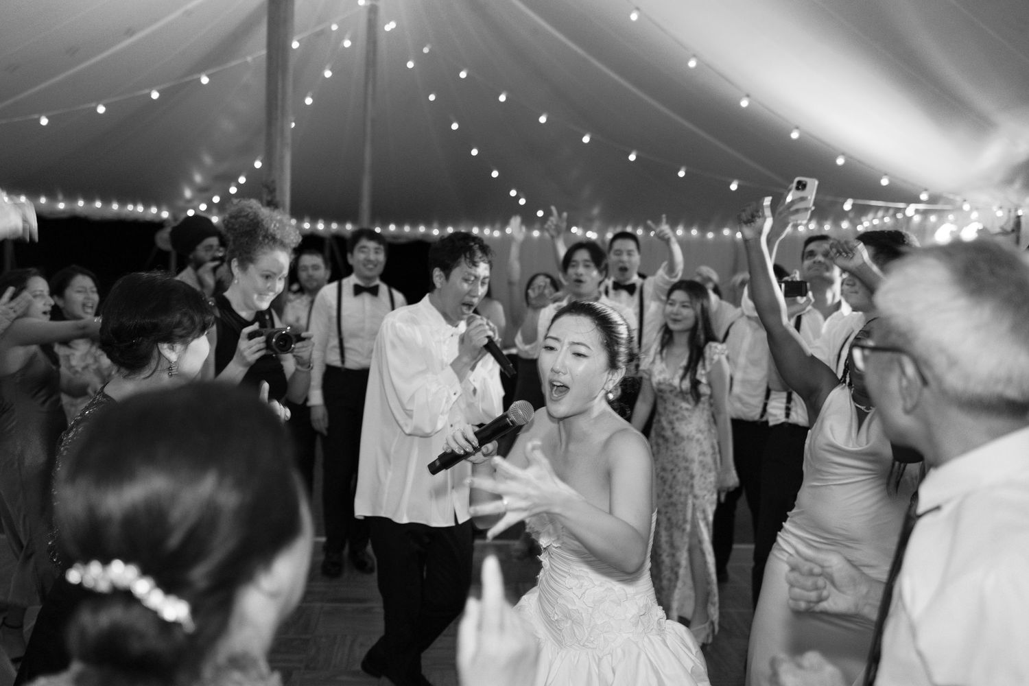 A black and white photo of a wedding reception dance floor with string lights overhead and guests dancing.