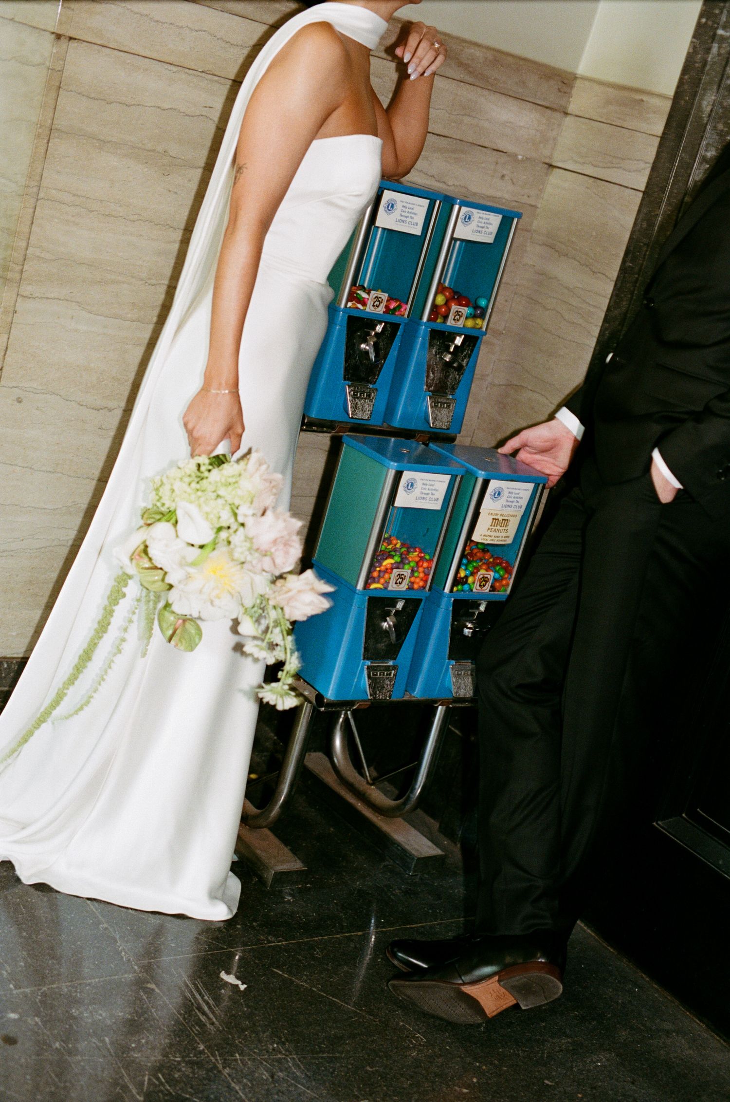 Bride in a strapless white gown holding a cascading white floral bouquet stands beside a groom in a black suit near blue gumball machines.