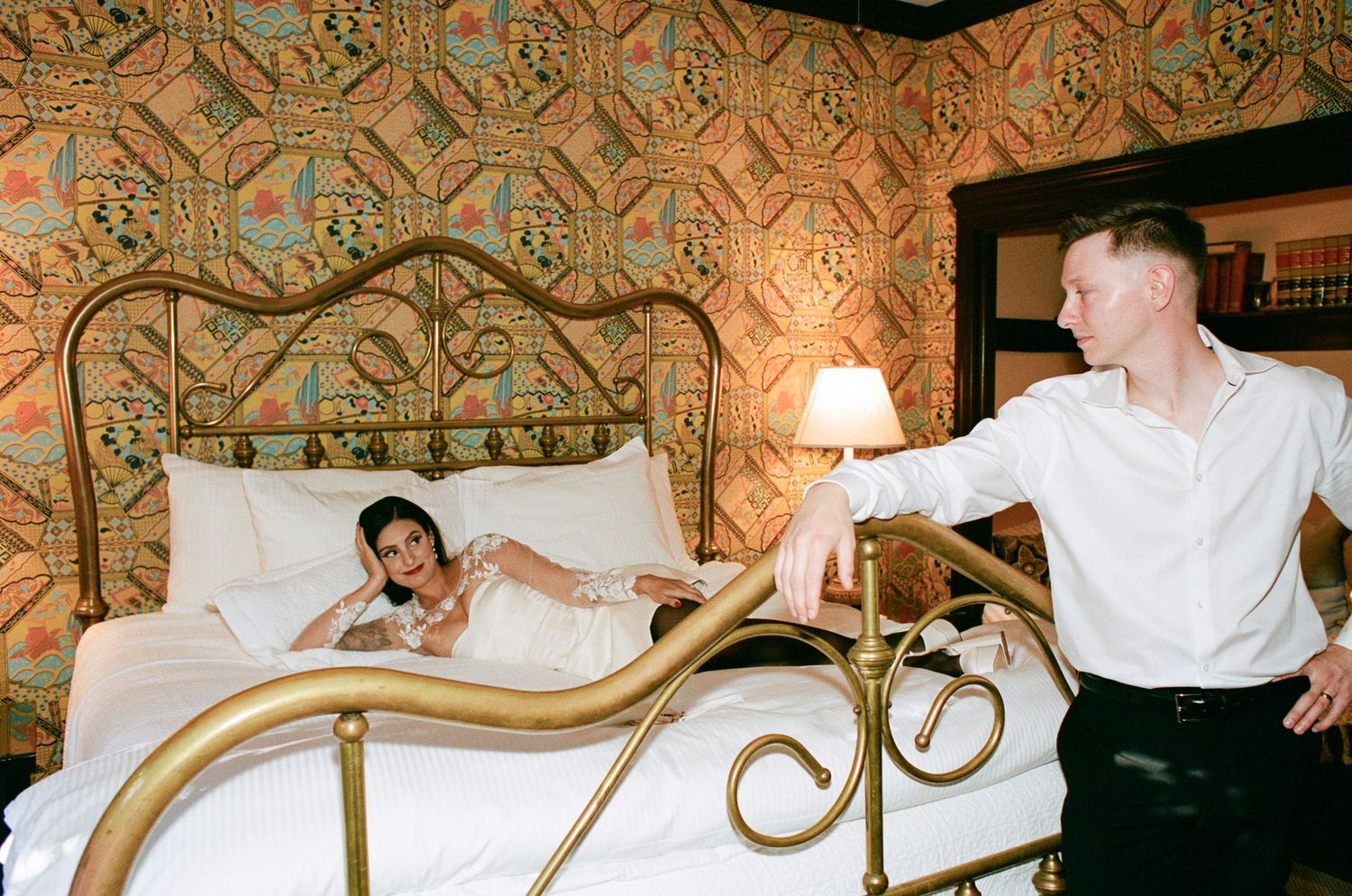 A man in a white shirt stands beside an ornate brass bed where a woman reclines, in a room with patterned gold wallpaper.
