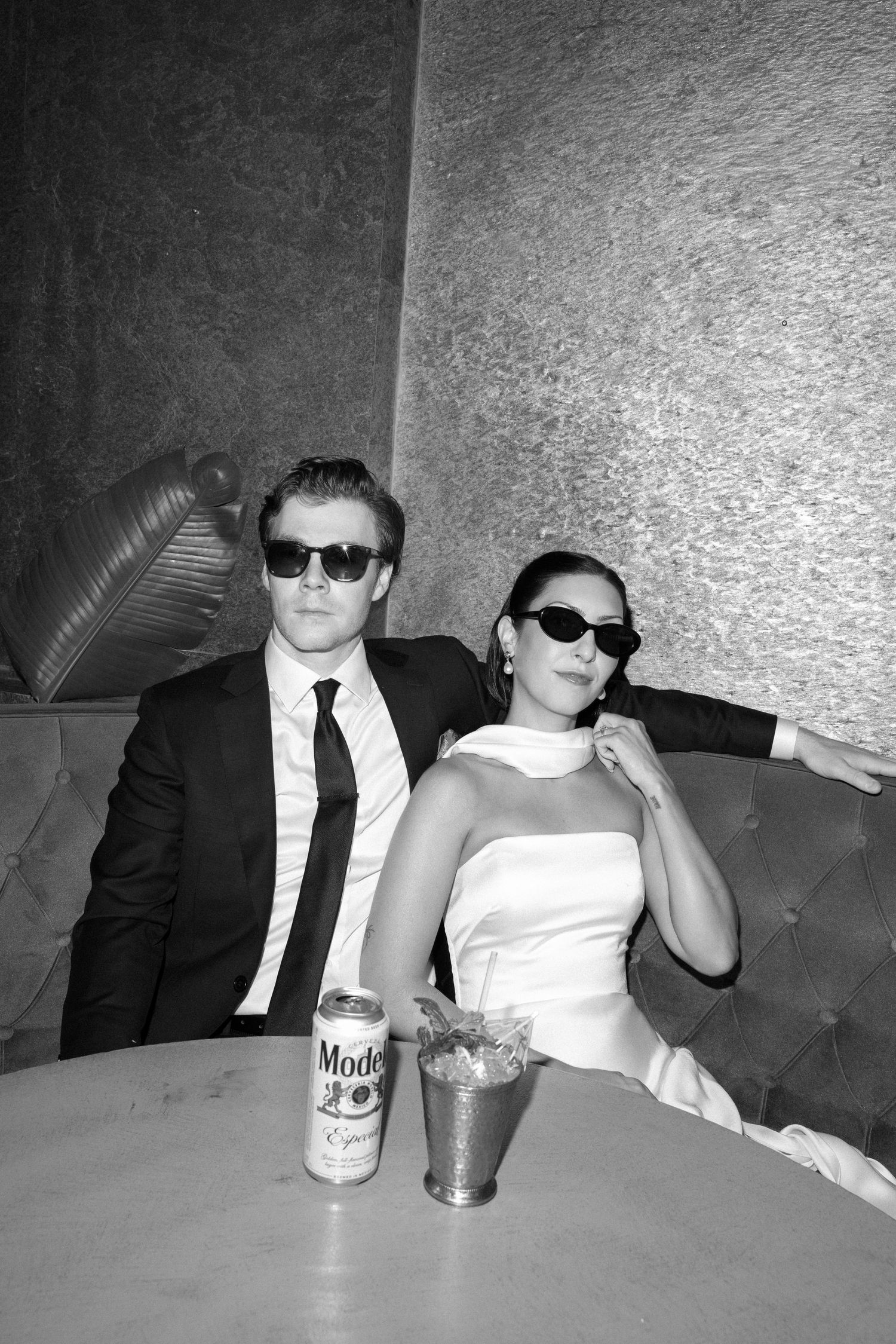 Black and white photo of a couple in sunglasses, he in a suit and tie, she in a white dress, seated in a tufted booth with a Modelo beer.