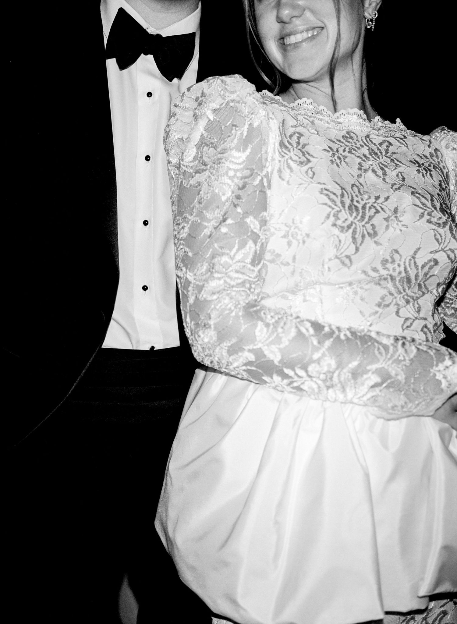 Black and white photo of a bride in lace wedding dress and groom in tuxedo at a formal wedding celebration.