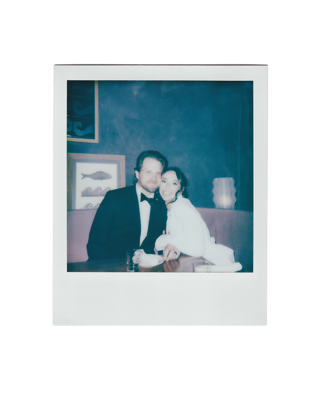 A couple poses for a Polaroid photo, man in tuxedo and woman in white fur, against a teal blue wall.
