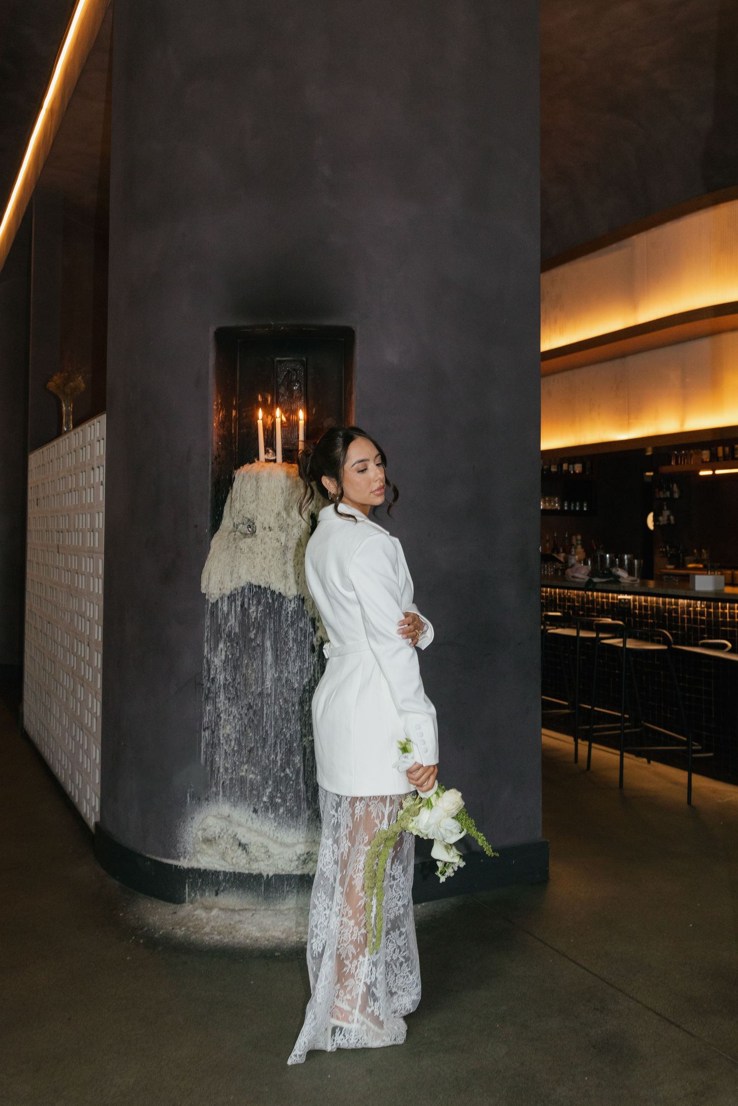 Bride in white blazer and lace skirt holding bouquet poses by fireplace in elegant dark-toned venue.
