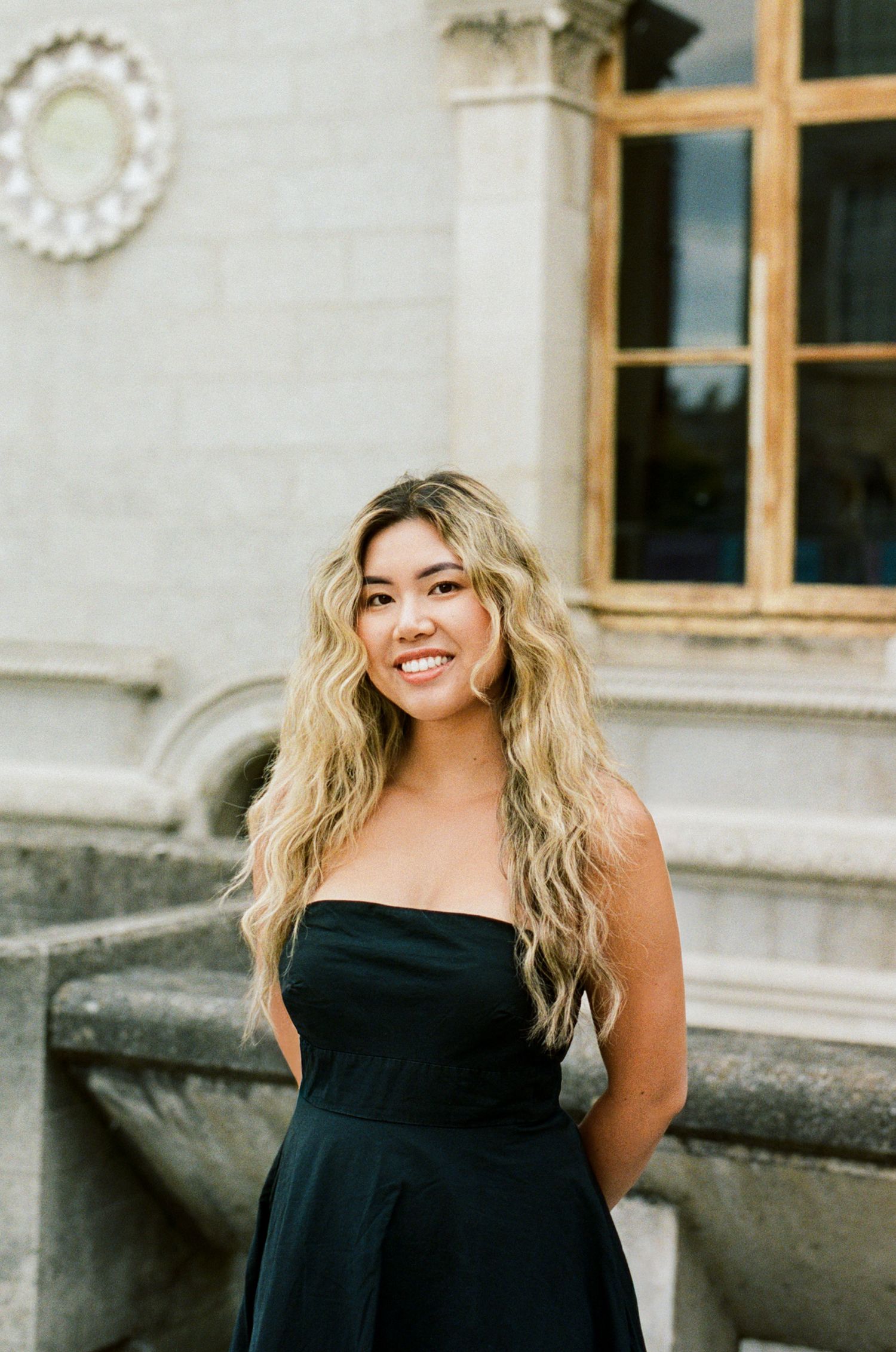 Young woman with long blonde wavy hair smiling in a black strapless dress, posing outdoors near a stone building.