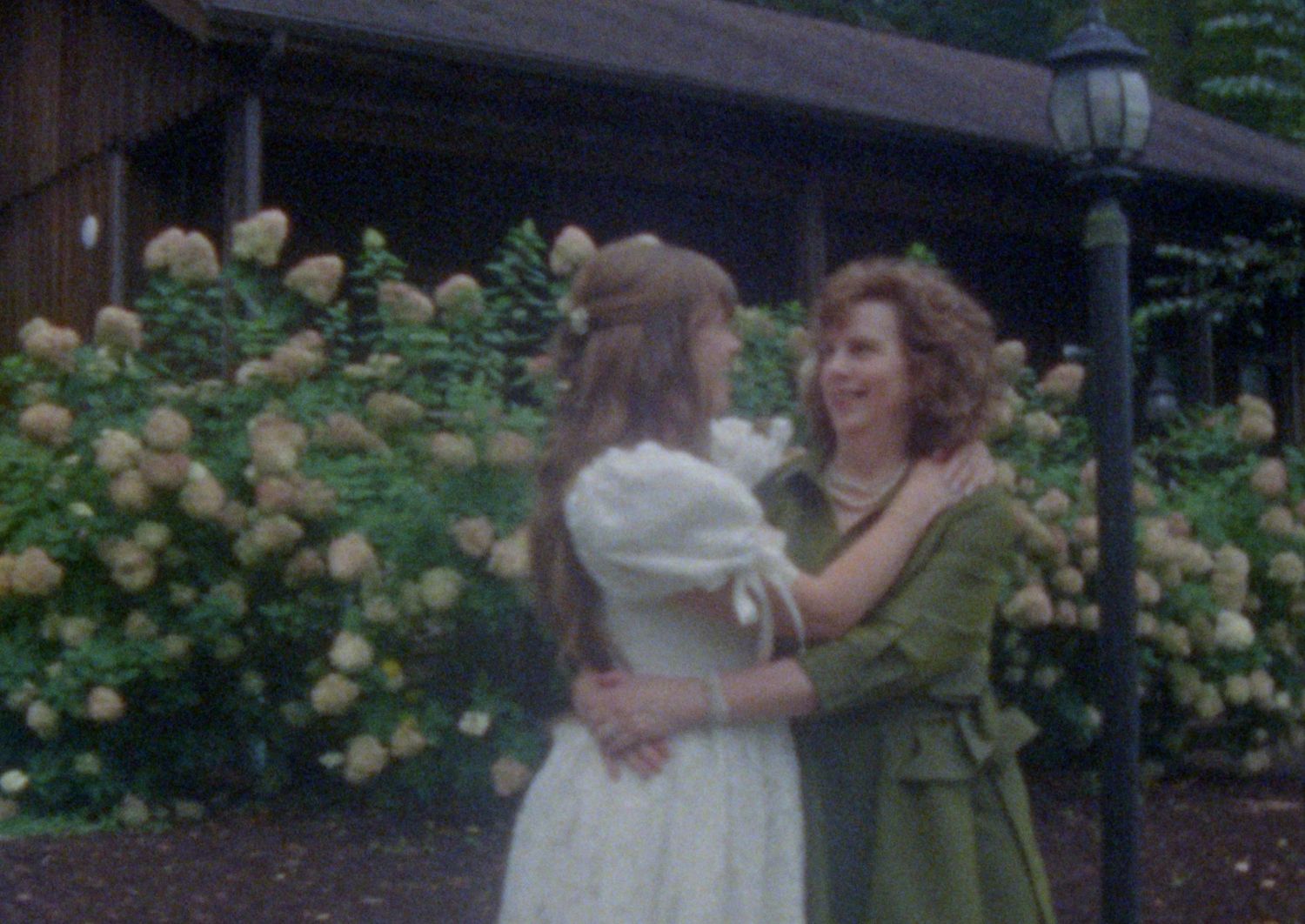 A couple in green and white dresses dances together in a garden filled with yellow roses.