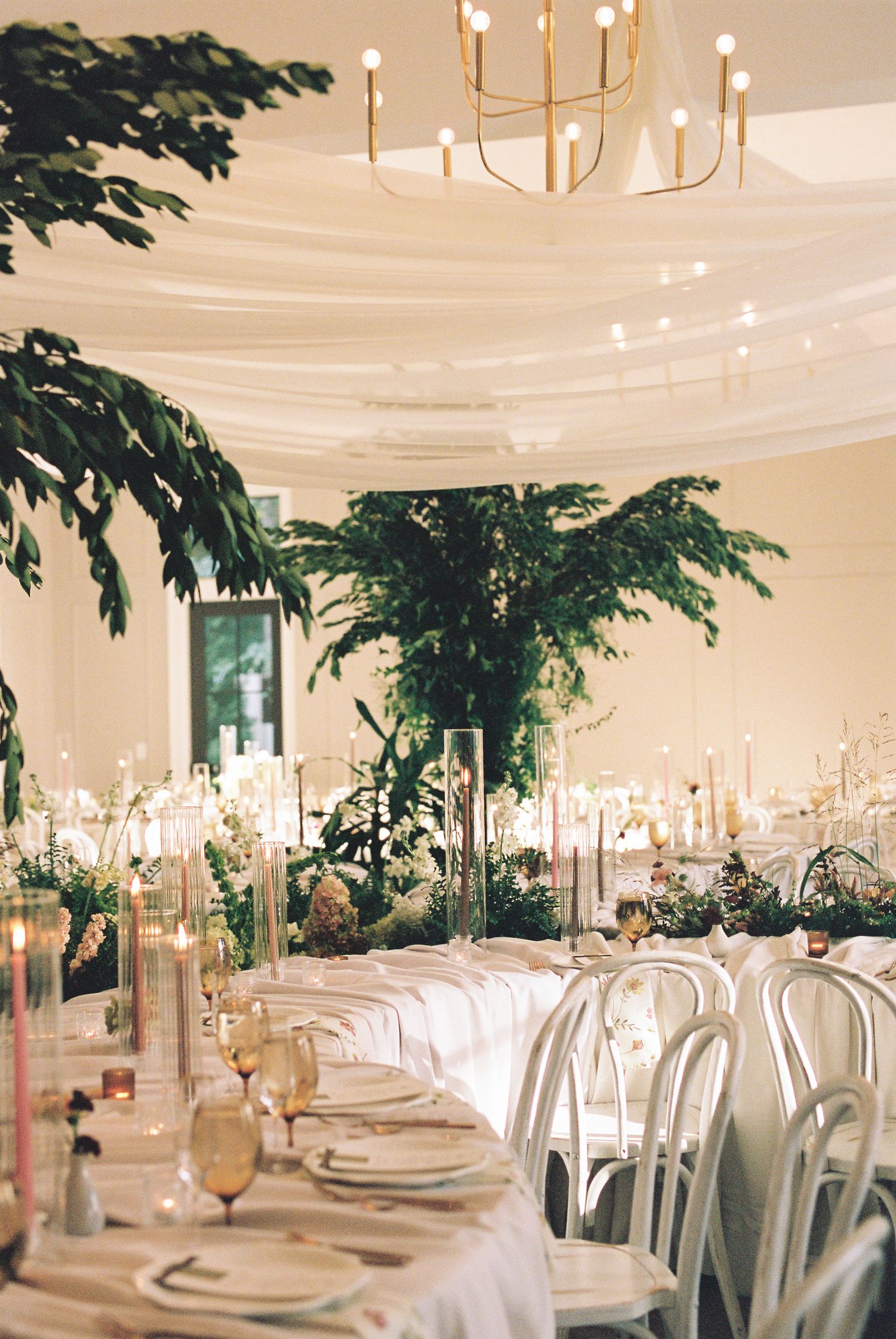 Elegant wedding reception with lush greenery, white draping, candlelight, and gold chandelier centerpiece.