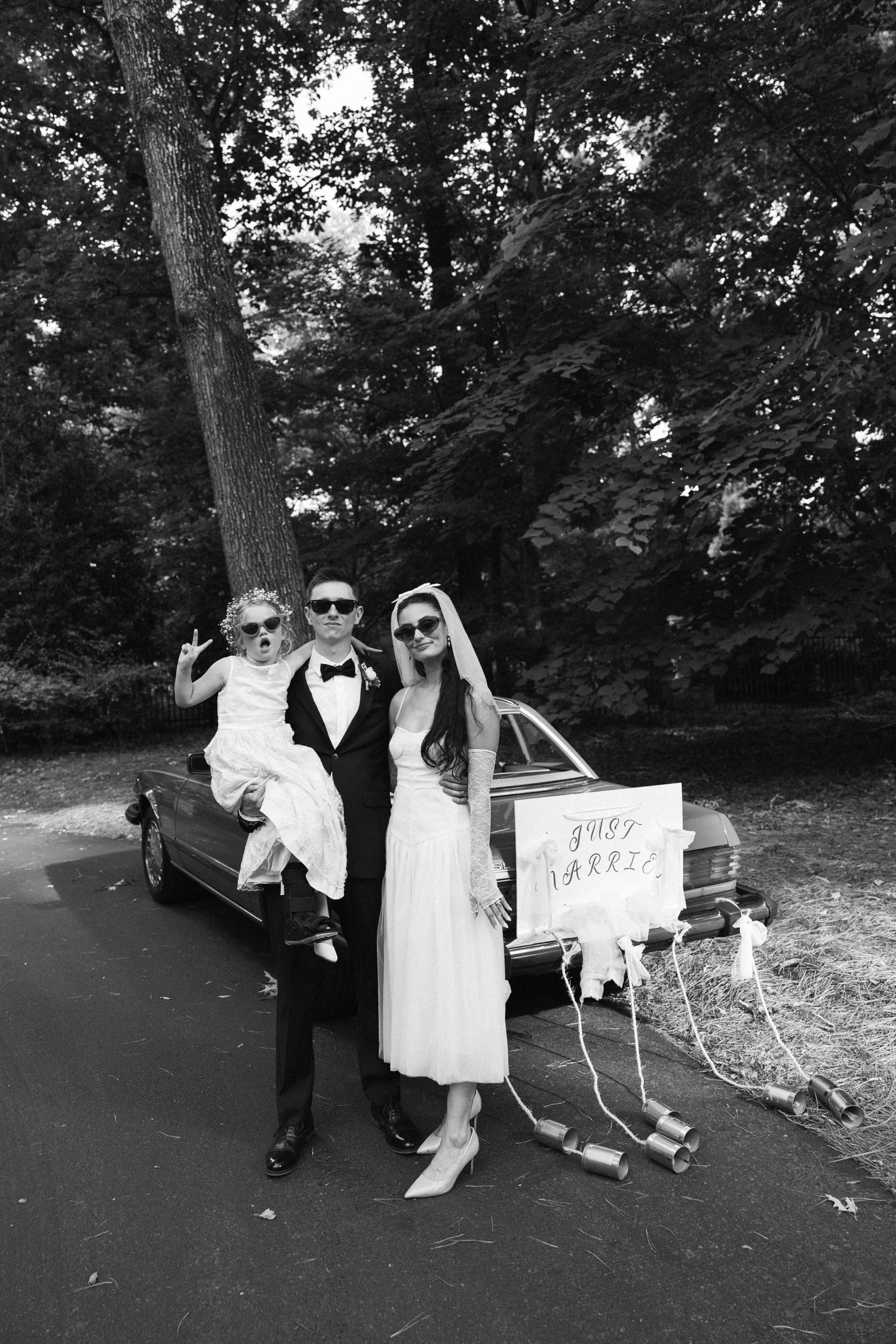 A stylish wedding party poses with a vintage car in a black and white photograph.