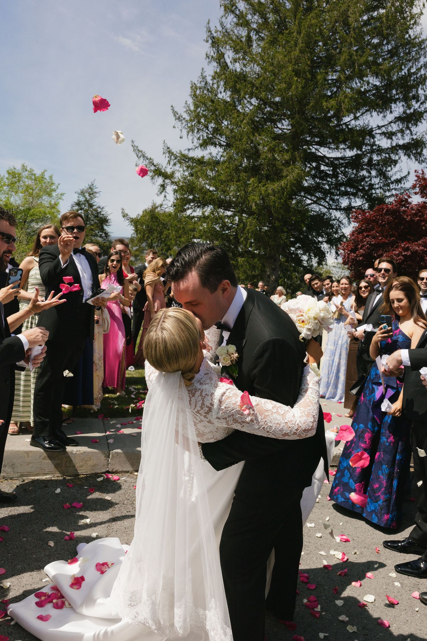 Bride and groom share first kiss as wedding guests toss rose petals outdoors on sunny day.