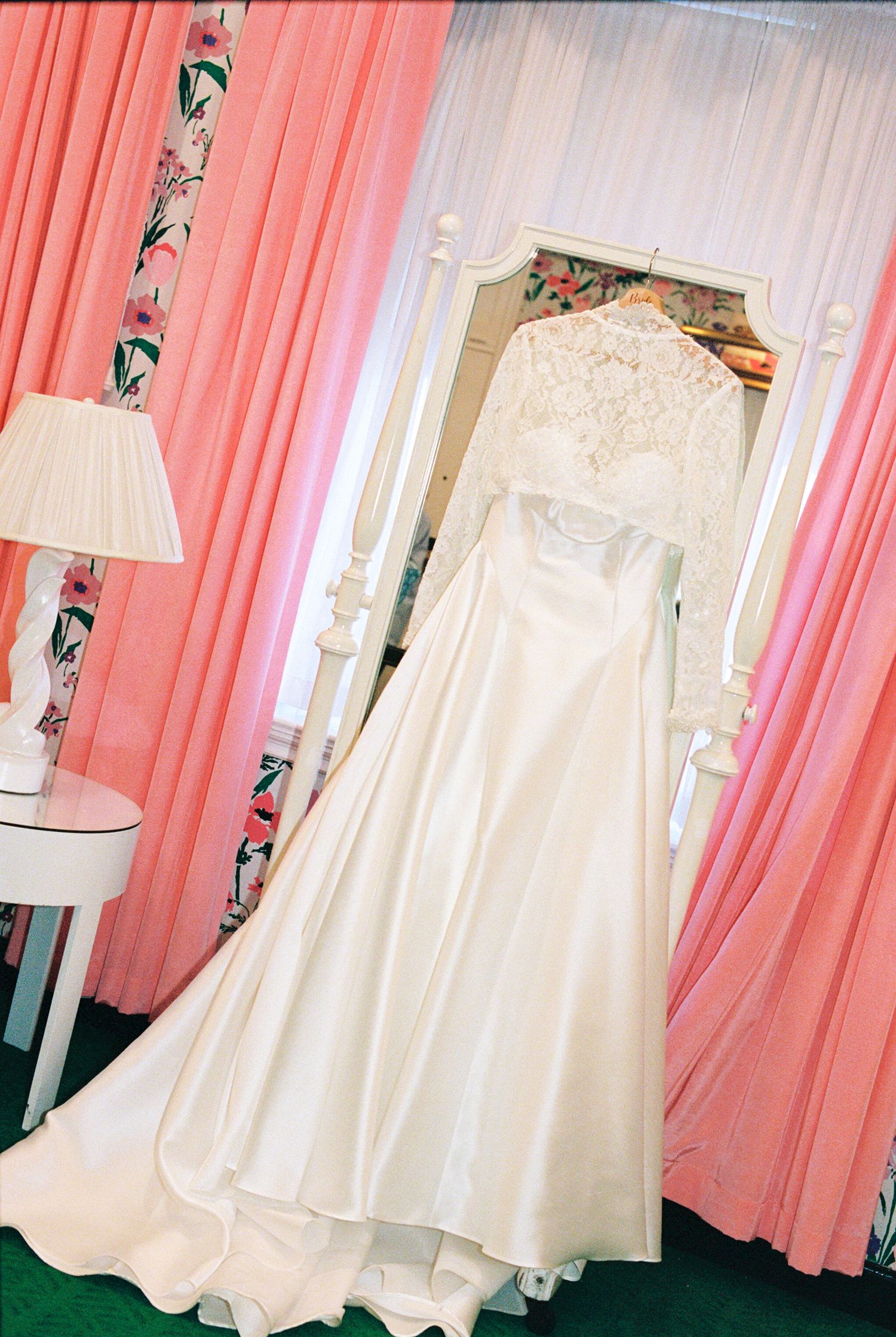 Elegant ivory wedding gown with lace bodice and satin skirt displayed on a hanger against pink curtains.