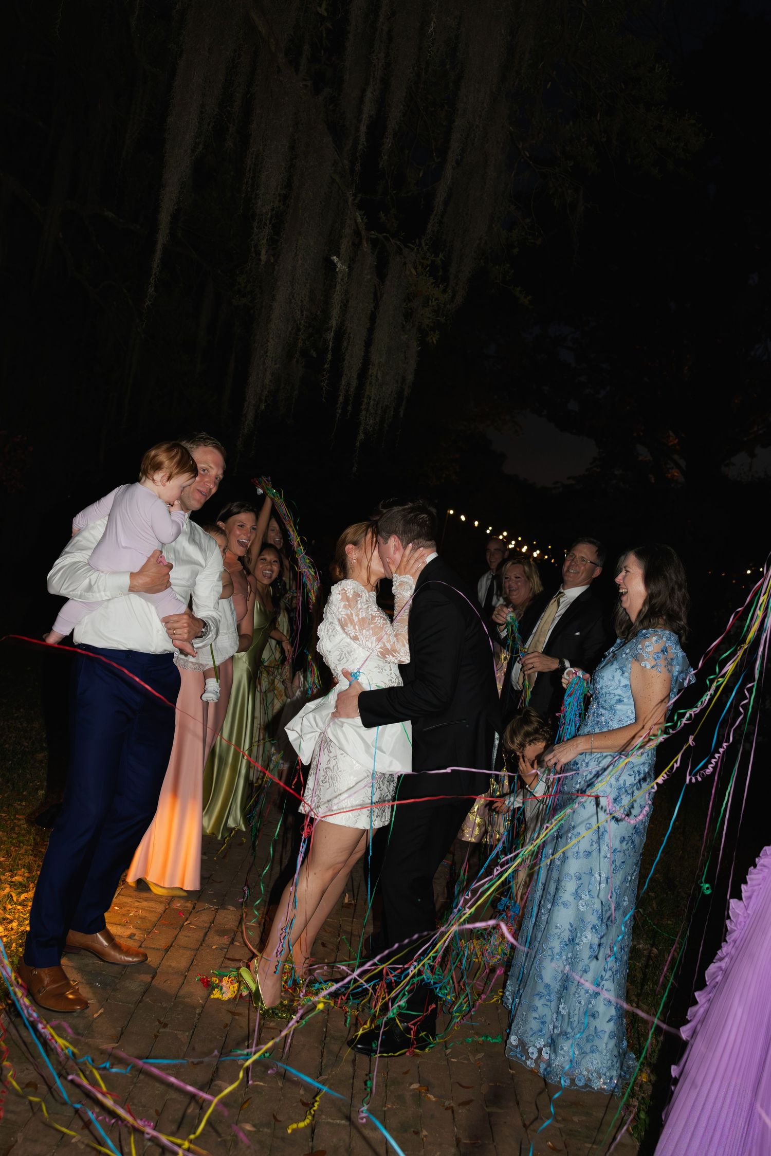 Wedding couple kissing surrounded by guests throwing colorful streamers outdoors at night celebration.