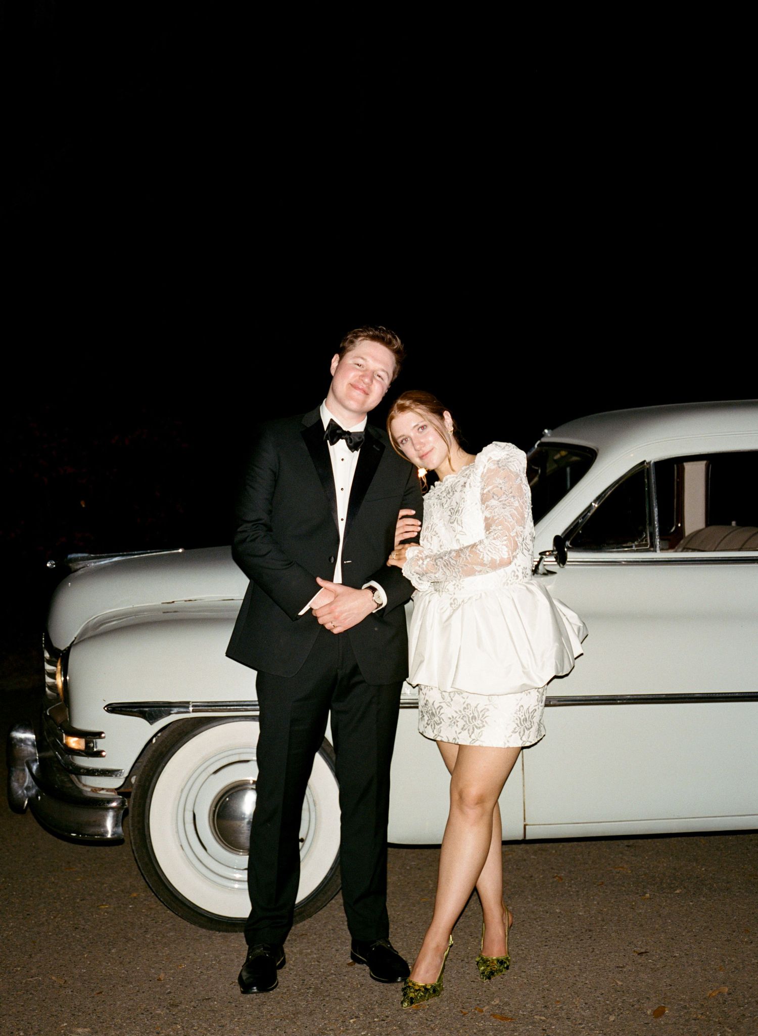 Couple in wedding attire posing by a vintage white car at night, groom in tuxedo and bride in short lace dress.