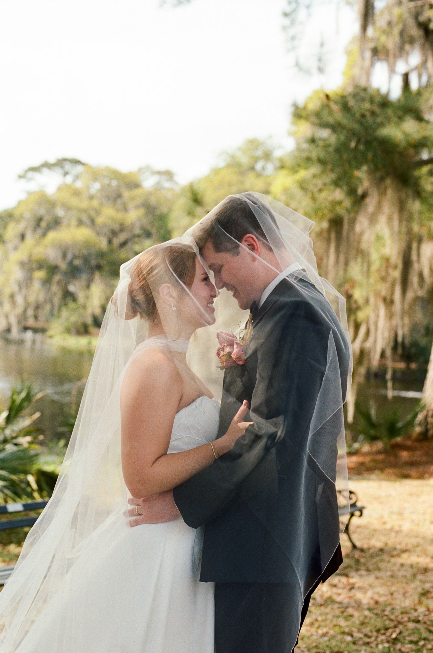Bride and groom share romantic moment under wedding veil outdoors near pond with Spanish moss trees.