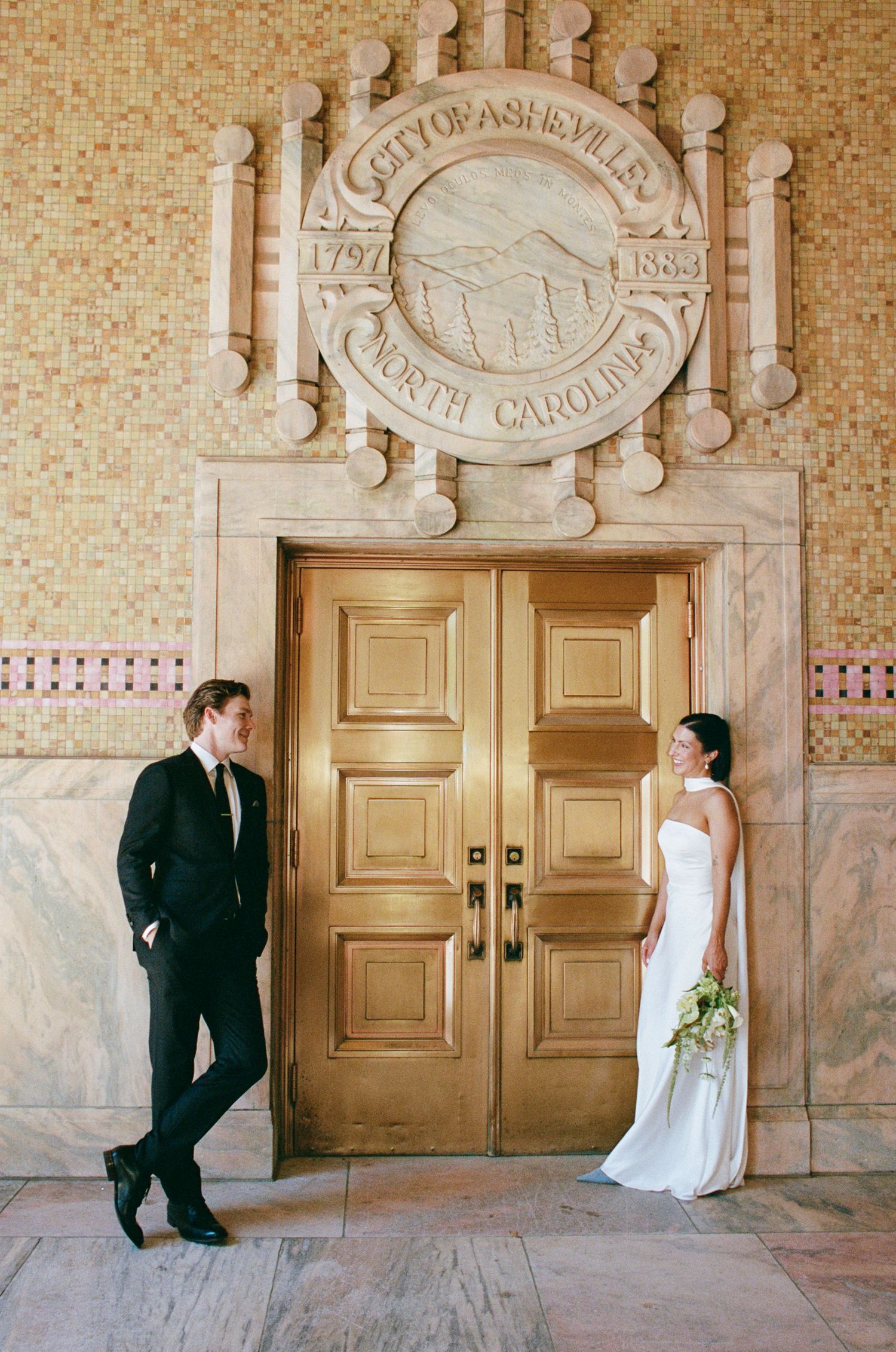 Bride in a white gown with bouquet and groom in a black suit stand beside golden doors under the City of Asheville seal, North Carolina.