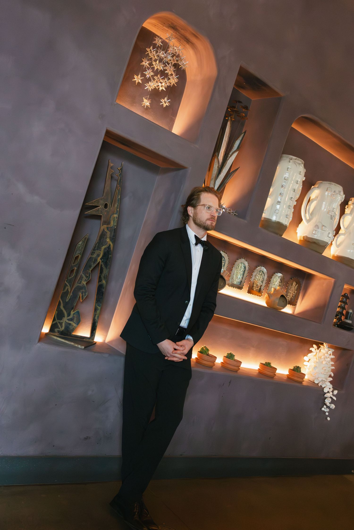 Man in black tuxedo standing by illuminated decorative shelves with artistic objects in elegant interior.