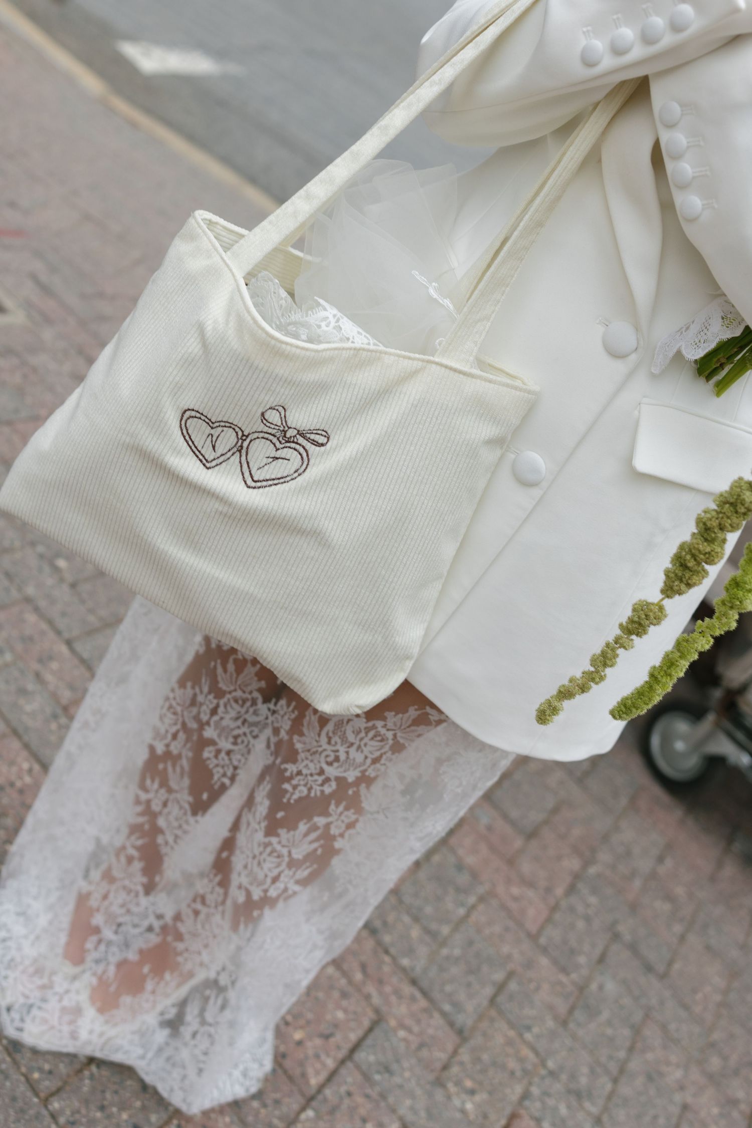 Elegant bridal accessories featuring an ivory tote bag with heart embroidery, white lace dress, and buttoned blazer on cobblestone.