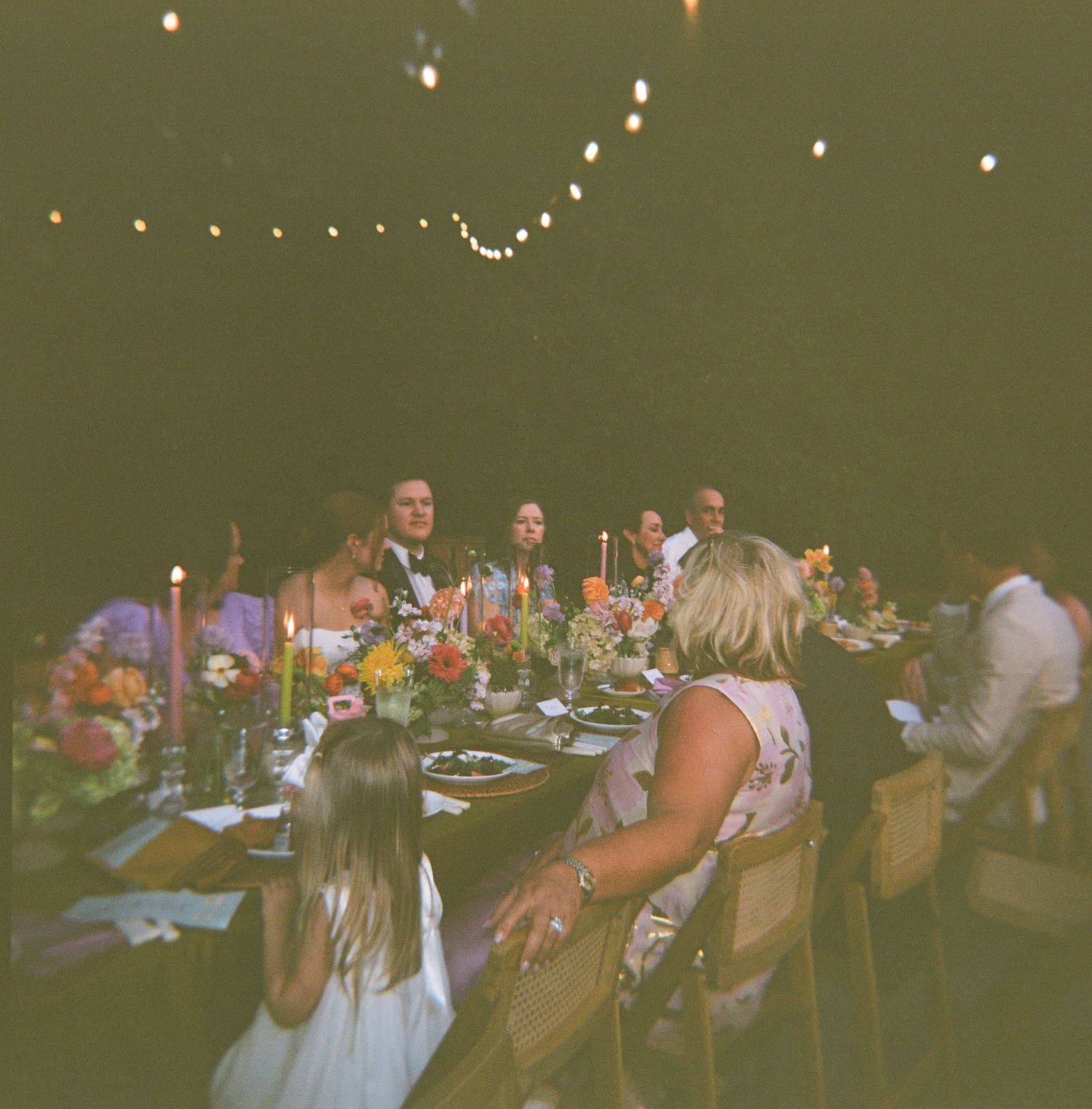 Outdoor wedding reception dinner at night with string lights, colorful floral centerpieces, and candles on long tables.