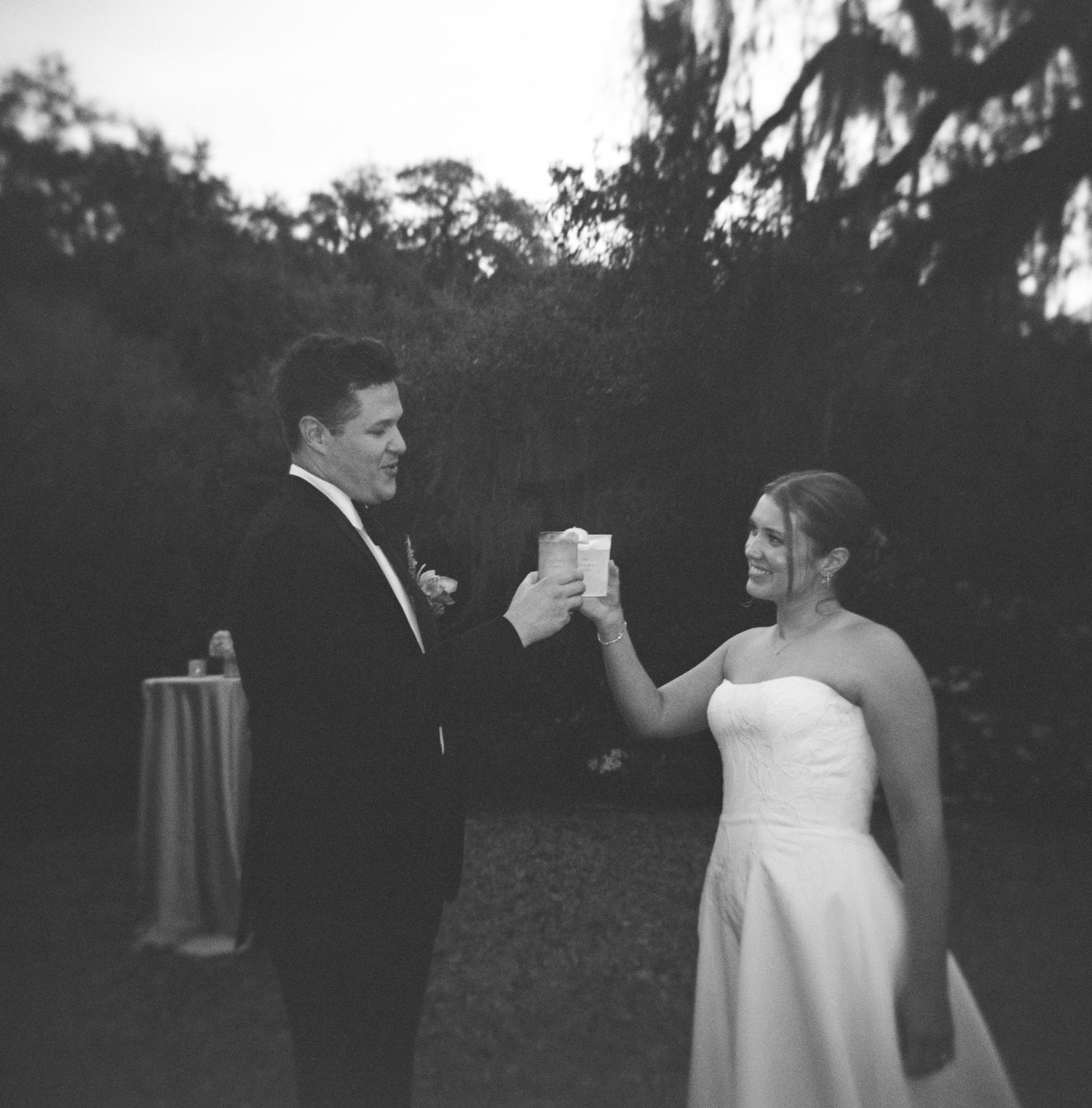 Black and white photo of bride and groom toasting with cups at outdoor wedding reception.