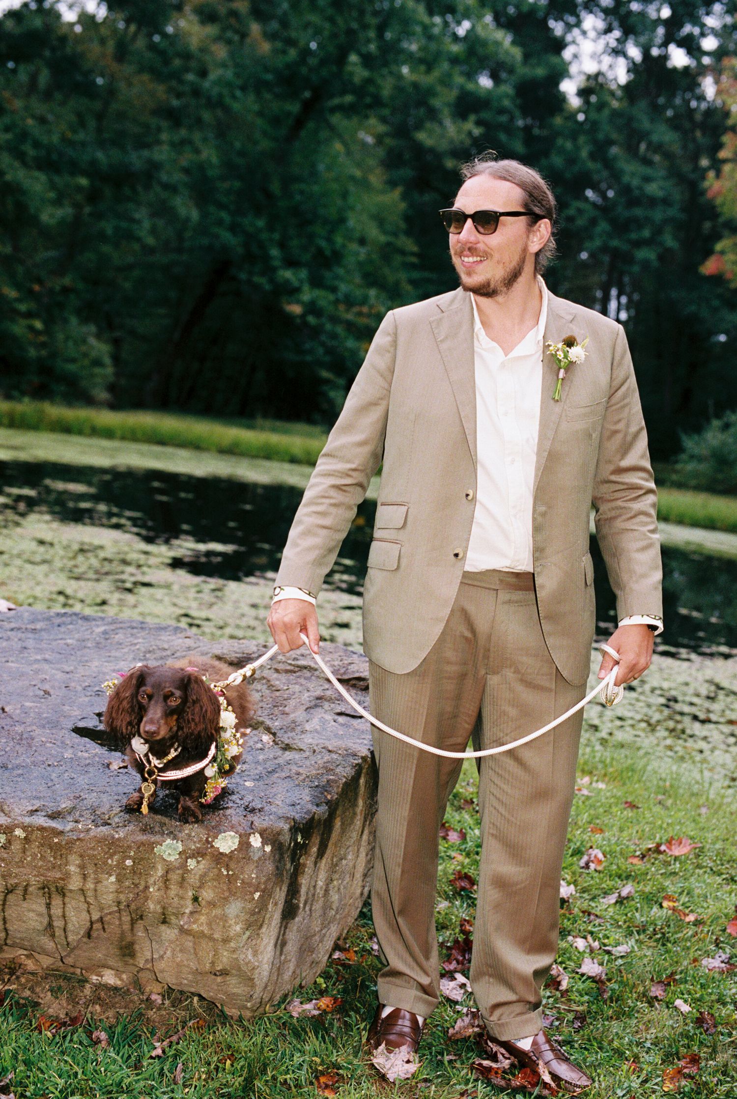 A person in a tan suit poses with a chocolate Labrador retriever dog by a stone wall near a pond.