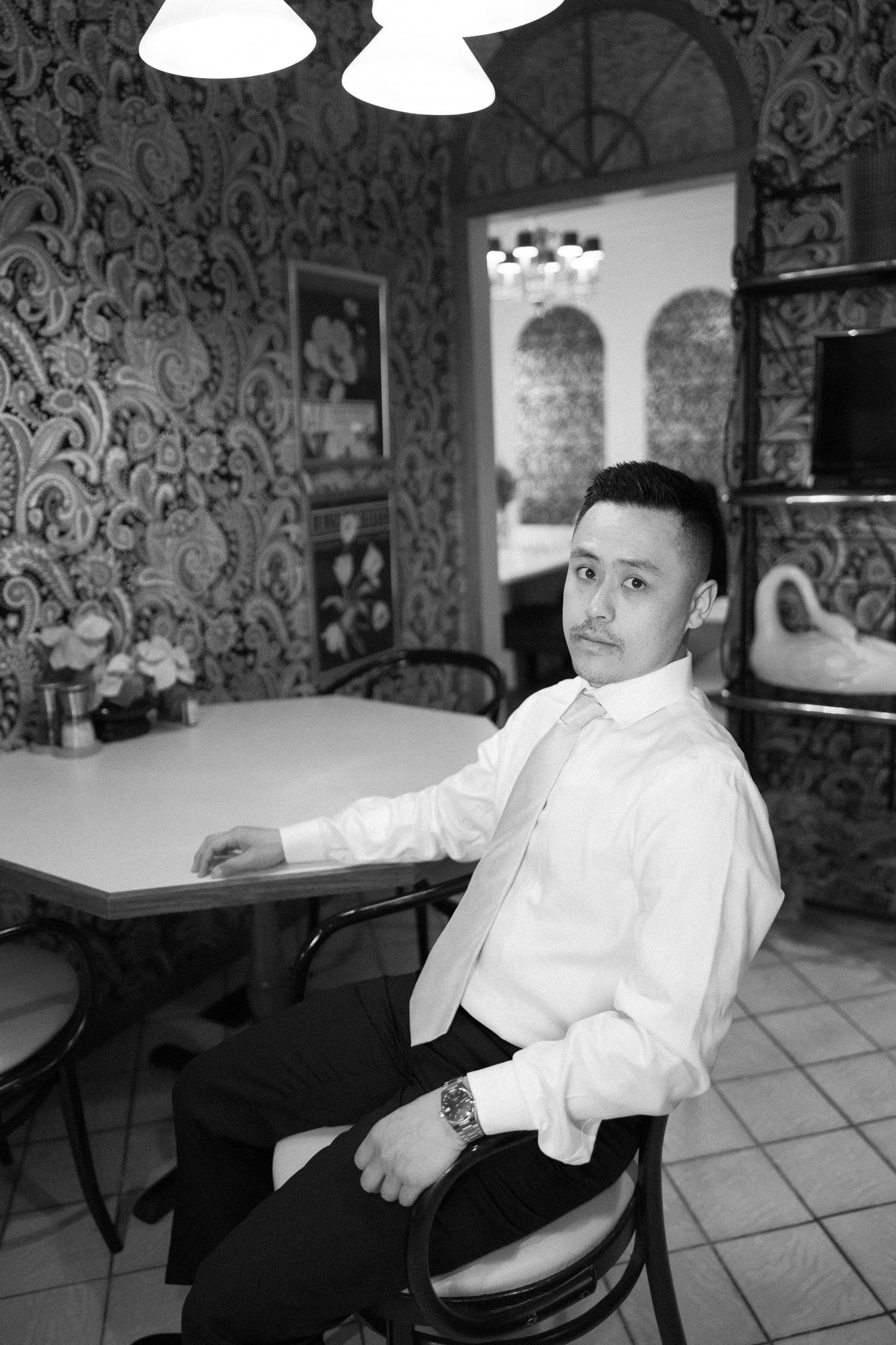 Black and white photo of someone in formal attire seated at a table in an elegant room with ornate wallpaper and lighting.