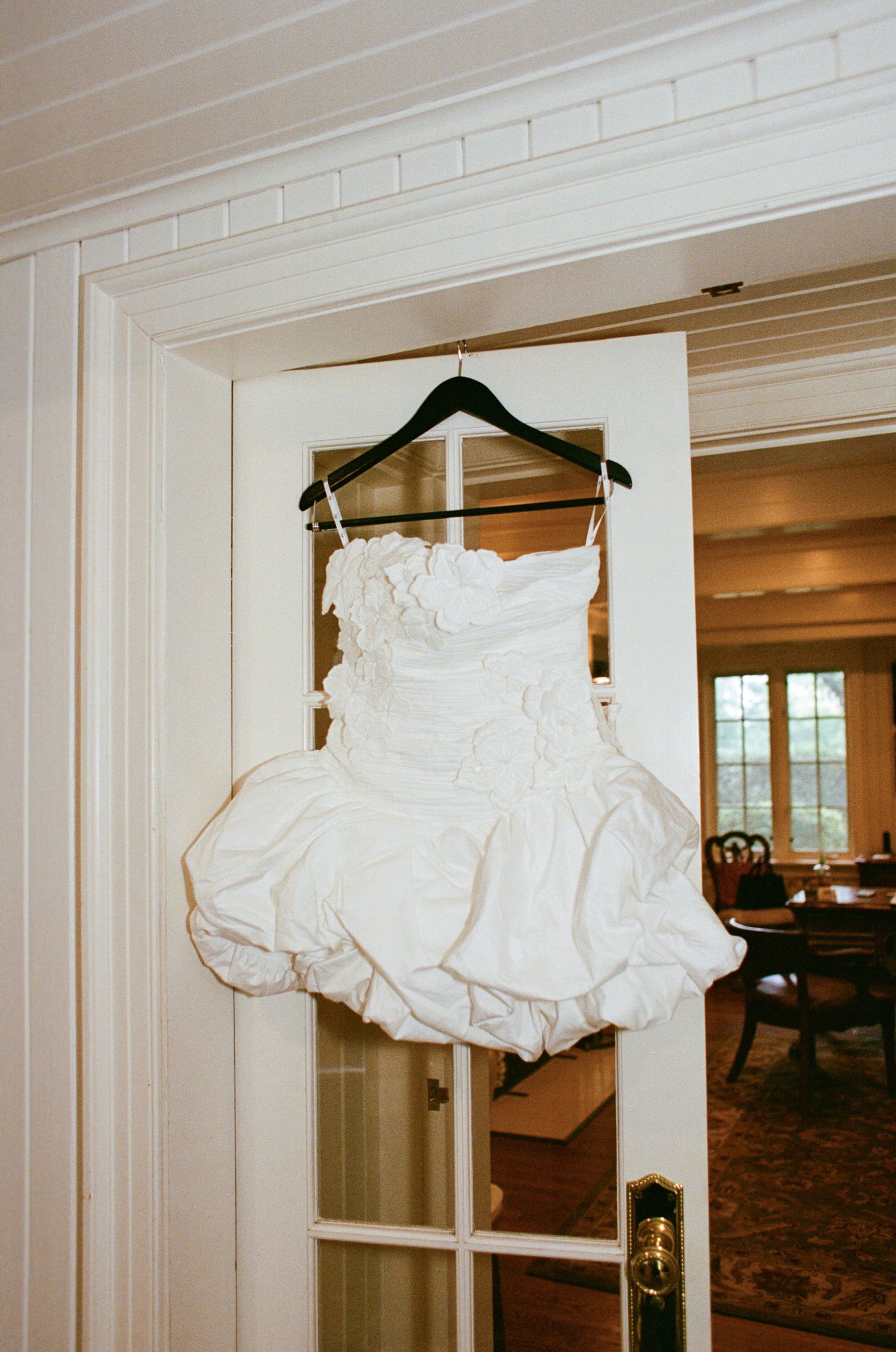 A white wedding dress with ruffled skirt hangs on a wooden hanger against a white doorframe in natural light.