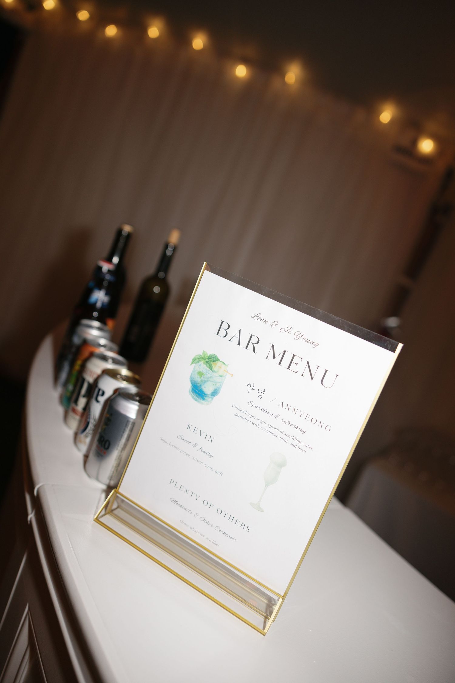 A bar menu sign displayed in a gold frame with string lights and liquor bottles in the background.
