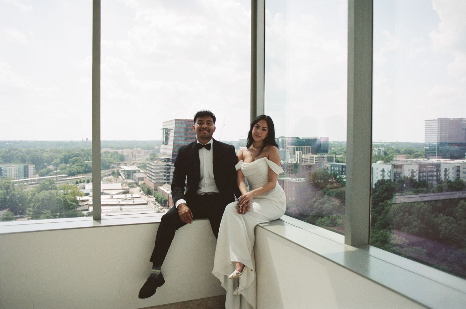 A couple sits together on a window ledge overlooking a cityscape view from a high-rise building.