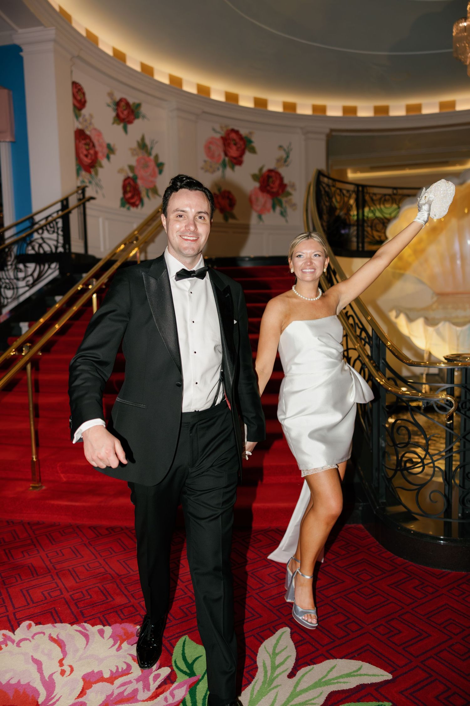 Elegant couple in formal attire posing on red carpet staircase, man in tuxedo and woman in white strapless dress.