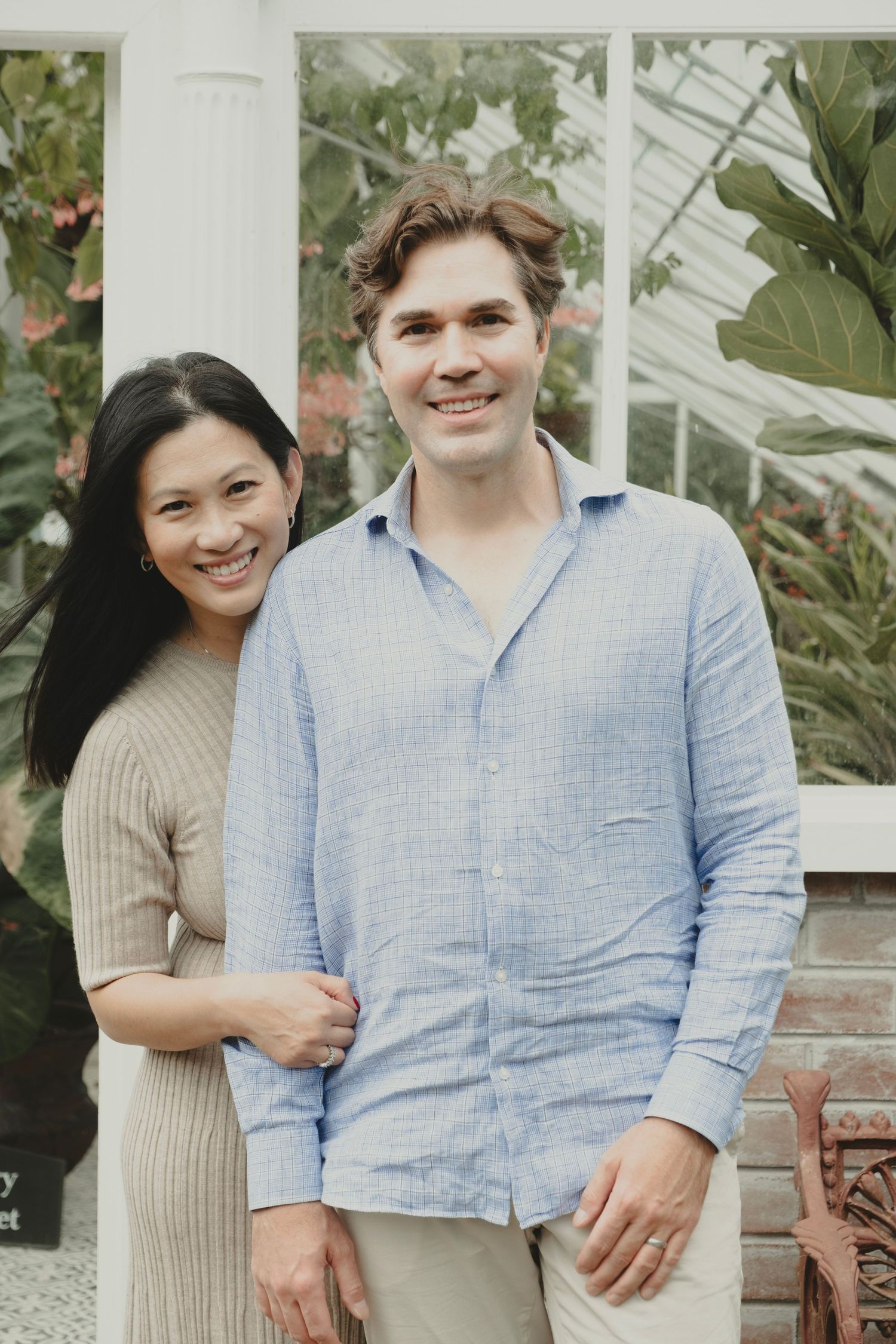 A couple poses together outdoors in casual attire with a beige sweater and blue linen shirt against a botanical background. Mincher gardens