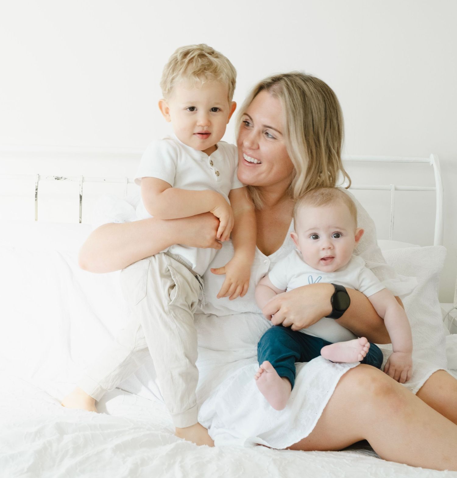 Mother in white clothing sits on bed playing with two young children surrounded by white bedding. auckland