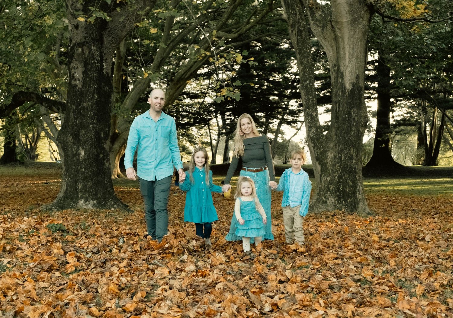 A family in matching blue outfits walks together through autumn leaves scattered under tall trees in a park. Cornwall park auckland