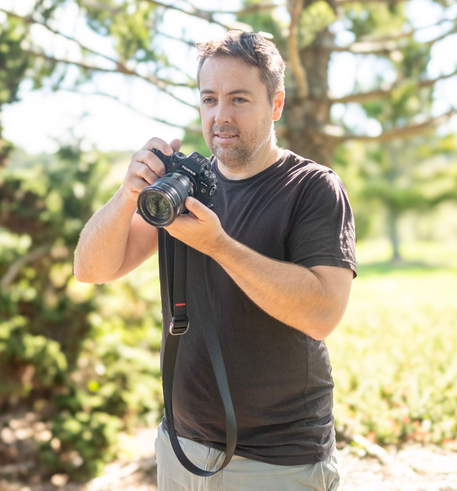A photographer wearing a black t-shirt holds a DSLR camera outdoors in a natural setting with trees in the background. Auckland Botanic
