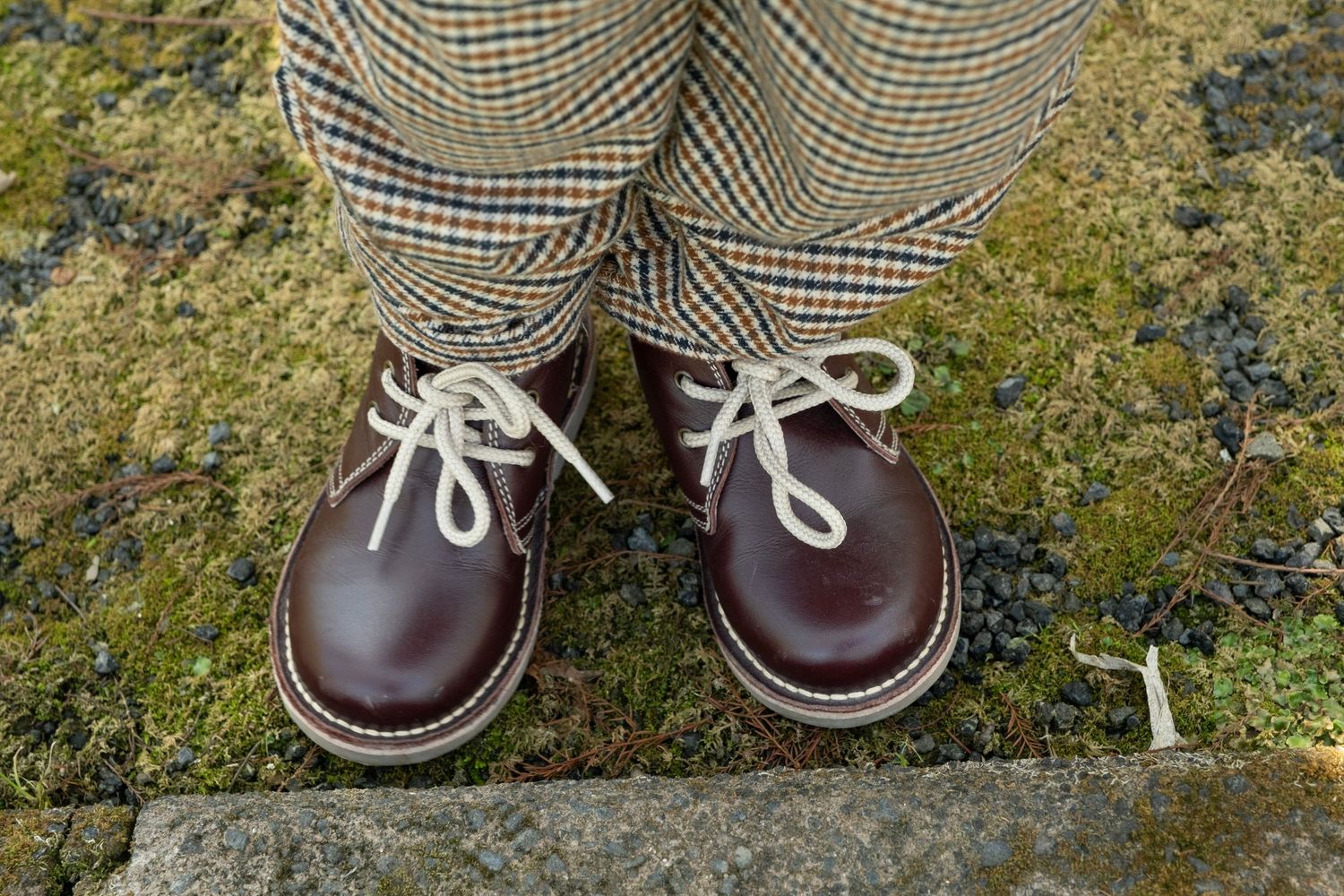 A pair of brown leather oxfords with white laces worn with plaid trousers on a mossy stone surface. daydreampics auckland